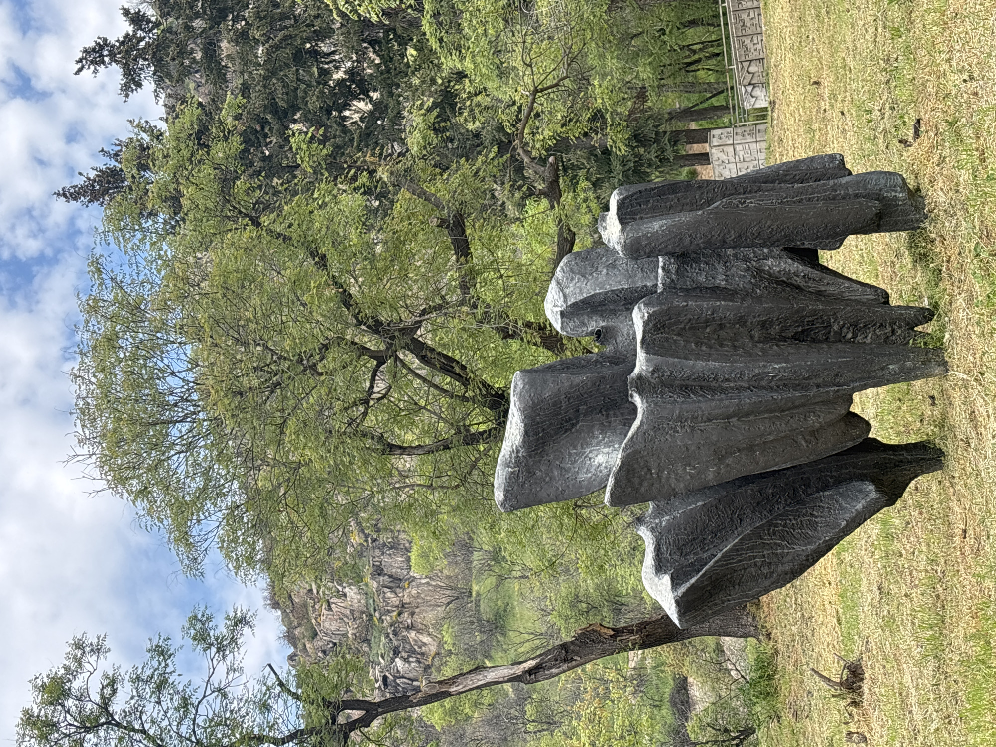 A modern, dark-colored abstract sculpture, possibly made of stone or metal, dominates the right side of the frame. Its surface is rough and textured, featuring a series of stacked, organic, and wave-like forms. The sculpture is set upon a ground covered with dry, yellowish-green grass.
Behind the sculpture and extending across the mid-ground are several trees with varying shades of green foliage, suggesting a spring or summer setting. Some trees have lighter, more delicate leaves, while others appear denser and darker. In the upper left portion of the background, a large, rugged rocky cliff or hillside rises, partially obscured by more greenery.
A light-colored fence or wall with distinct, stylized patterns or geometric shapes, resembling abstract text or decorative symbols, is visible in the mid-ground behind the sculpture, towards the right. The sky in the upper part of the image is bright blue with scattered white, cumulus clouds, indicating fair weather and daytime.
The scene depicts a tranquil, natural environment, likely a park or sculpture garden, featuring contemporary art. There are no people visible. This setting is located in Plovdiv, Bulgaria.