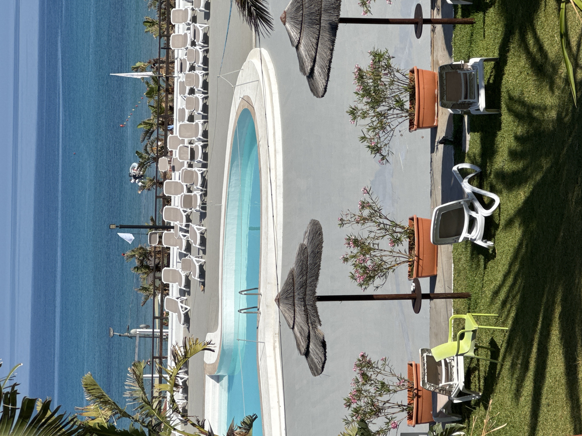 This image depicts a sunny coastal resort scene in Isola delle Femmine, Italy, observed from an elevated viewpoint. The foreground features a manicured green lawn with strong shadows of palm fronds. On the lawn, there are several white plastic lounge chairs and a single light green plastic chair. A prominent brown thatched umbrella with a dark central pole and a small circular shelf stands on the lawn, flanked by two terracotta planters holding flowering oleander plants with pink blossoms.
Beyond the lawn and planters, a gray paved area surrounds a kidney-shaped swimming pool filled with light blue water, bordered by white coping and equipped with metal ladders. Further back on the gray paving, numerous white and gray lounge chairs are arranged in rows. A dark wooden railing separates this area from the immediate coastline.
The midground reveals palm trees and other green foliage behind the railing. In the background, the Mediterranean Sea extends under a clear blue sky, displaying various shades of blue. A small white inflatable boat is anchored near the shore, and a line of colorful buoys marks an area in the water. A white flag on a tall pole is visible near the water's edge. No people are present in the scene.