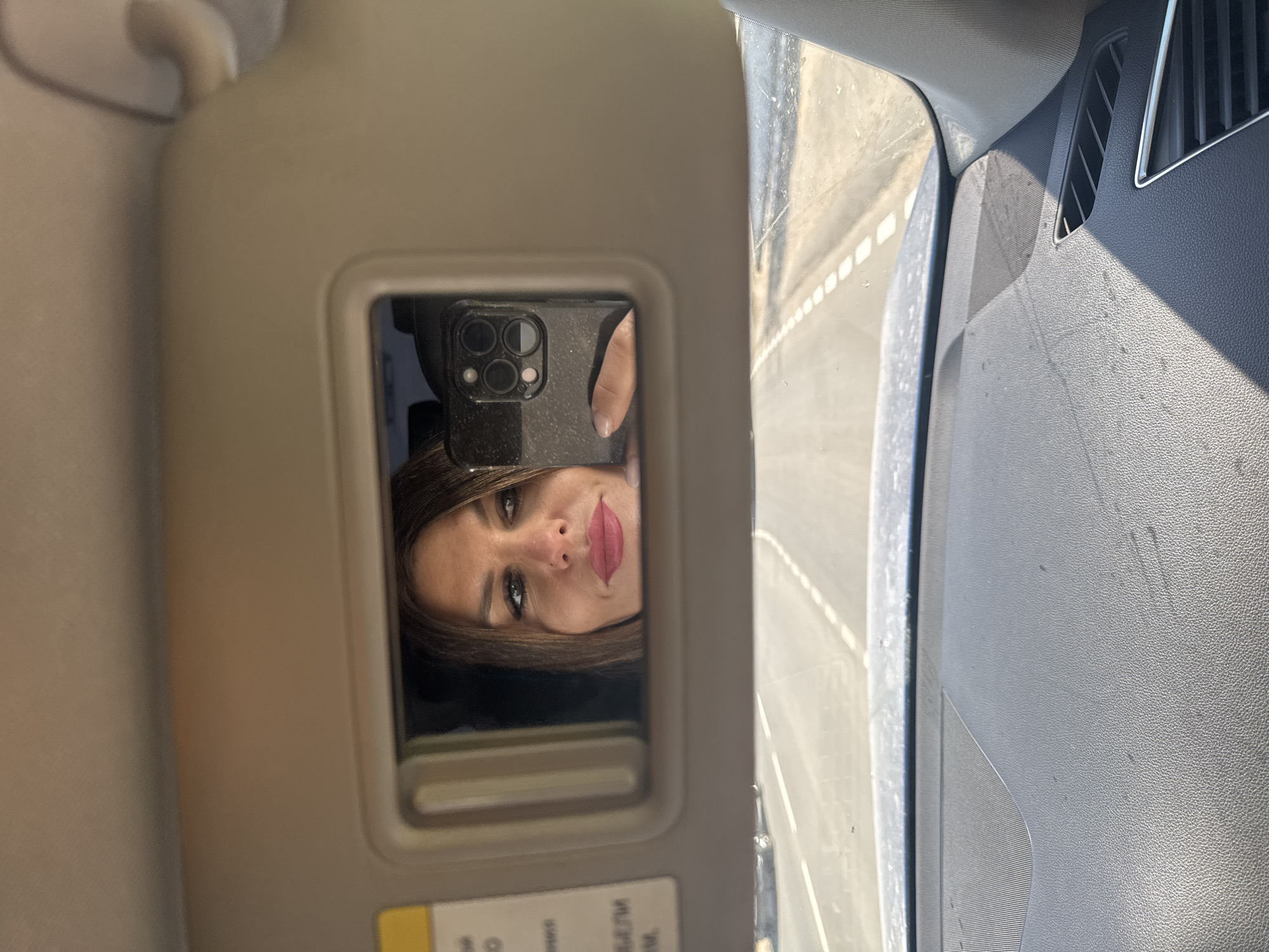 A woman is taking a selfie in a car. The image is reflected in the mirror of the sun visor. Outside the window is a road. There is no information that suggests the location is Chaikovsky, Russia.