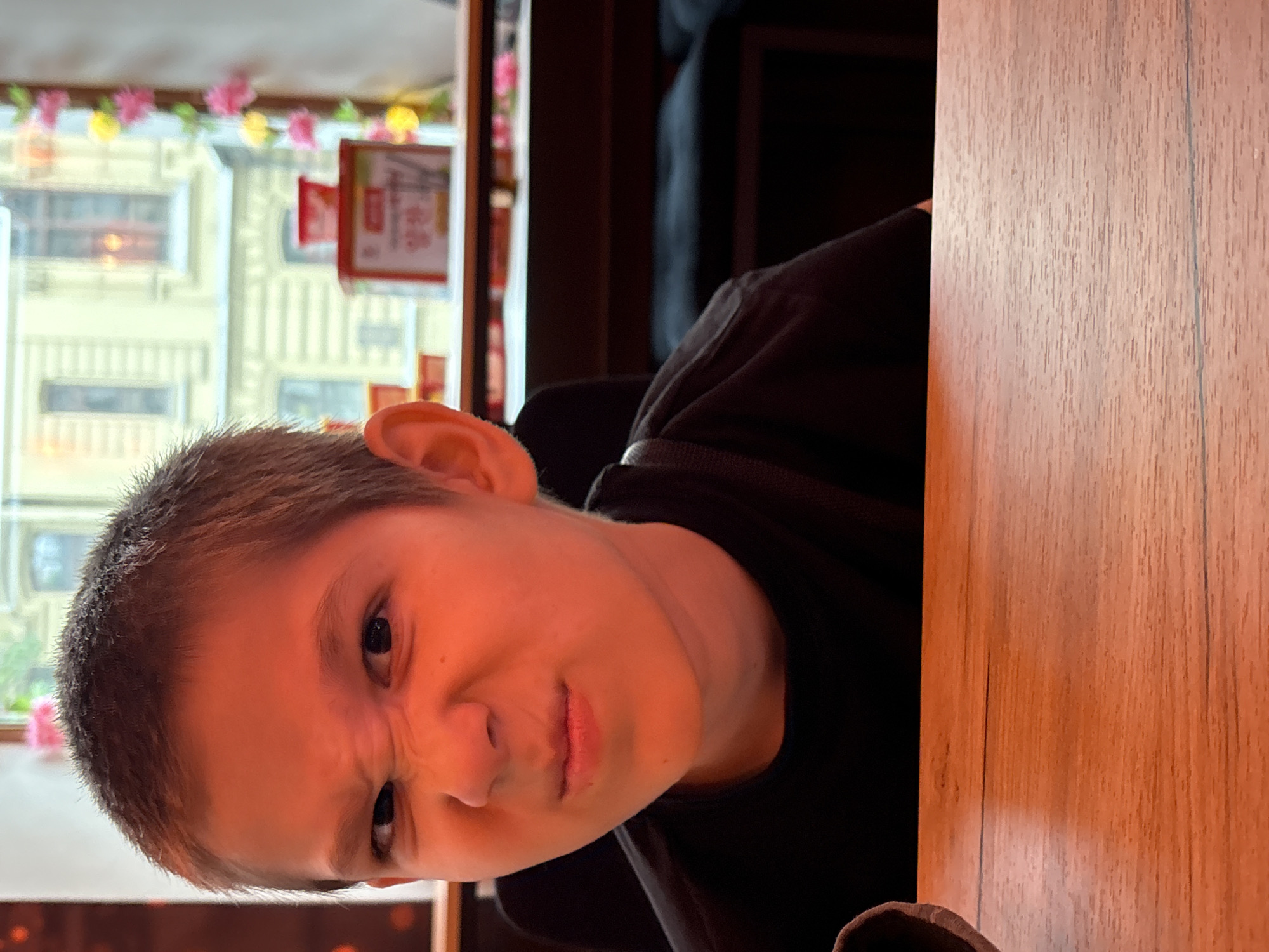 A young boy with short brown hair is pictured from the chest up, looking directly at the camera with a puckered and wrinkled expression. He is wearing a black t-shirt and is seated at a wooden table. Behind him, a window shows a blurred view of a building with multiple windows, possibly in an urban setting. Decorative pink and yellow flowers are strung along the window frame. A red box with some writing is visible on a shelf near the window. The lighting in the image is warm, casting an orange hue on the boy's face. The setting appears to be an indoor establishment with wooden furniture and dark seating in the background.