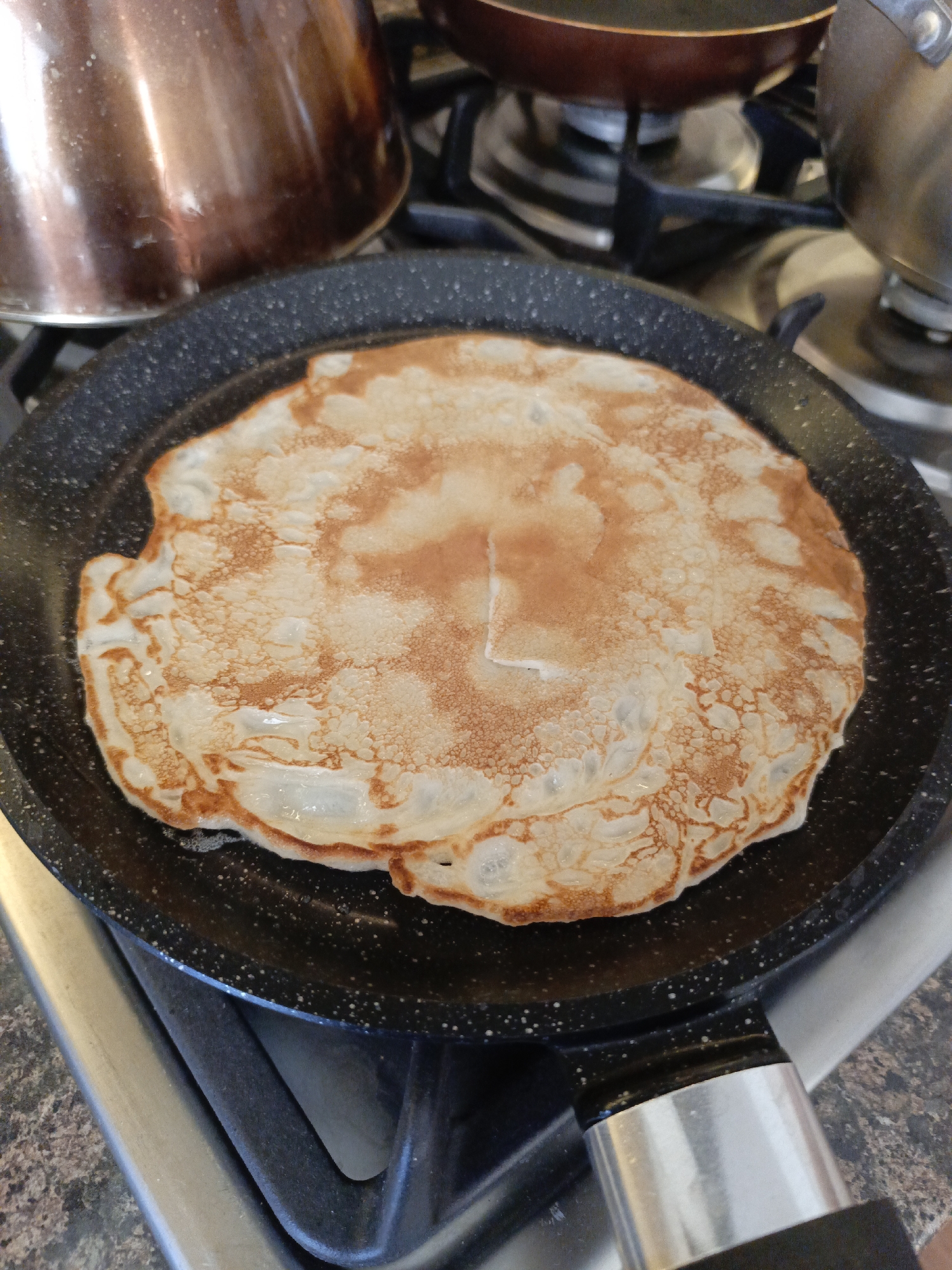 The image depicts a pancake cooking in a black frying pan on a gas stove. A copper-colored pot is visible in the background to the left, and a smaller frying pan and a pot are visible in the background to the right. The stove appears to be located on a kitchen countertop. The exact location in the city of Chaikovsky, Russia is not discernible from the image.