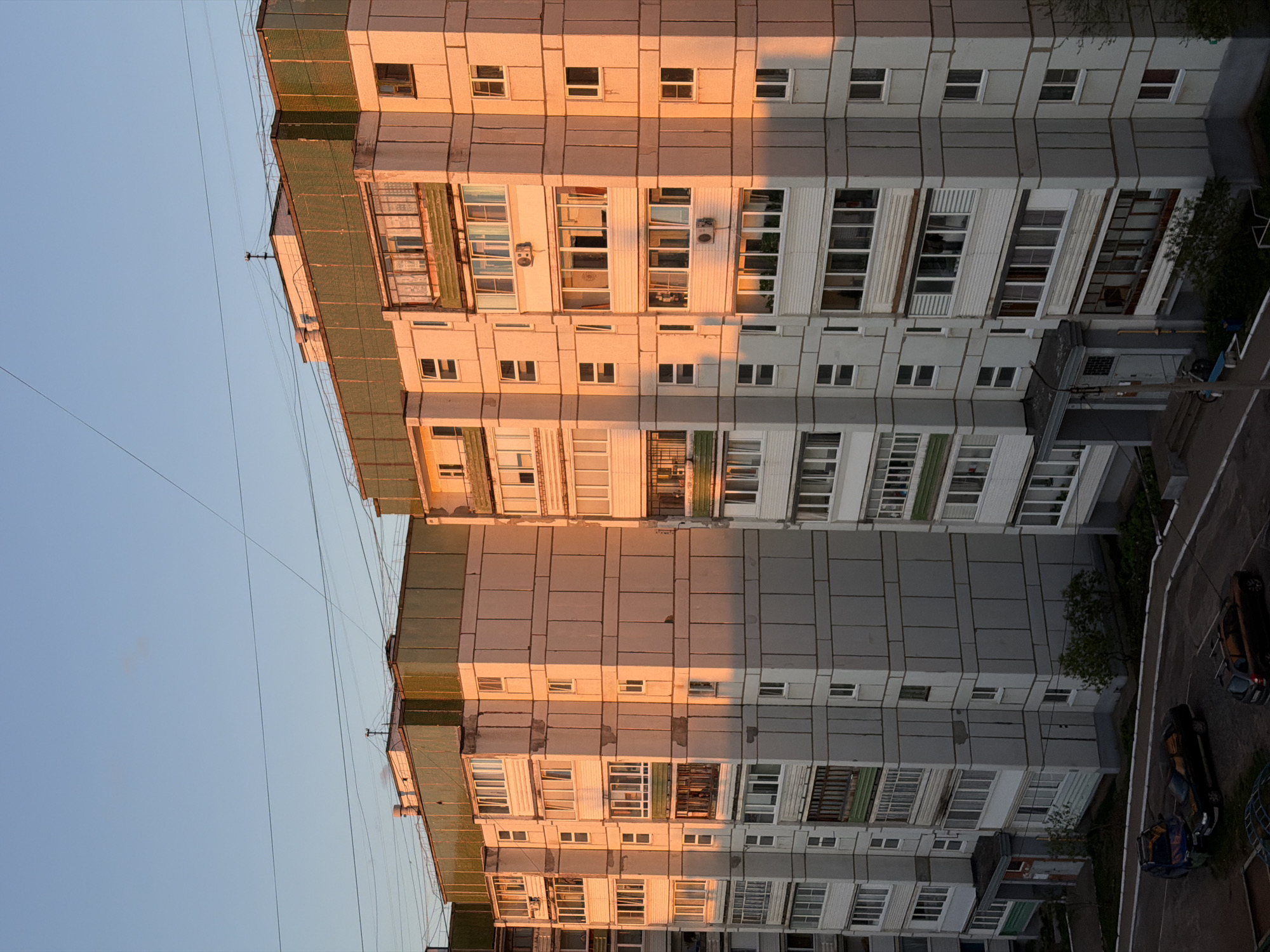 This image, likely taken in the city of Chaikovsky, Russia, features two multi-story apartment buildings. The buildings are constructed of concrete panels. The facade is divided into sections, some sections are painted in a light beige color that reflects the orange light of the setting sun, and others are painted grey. There are numerous rectangular windows on each floor. Some of the windows have air conditioning units. Between the two buildings, the sky is visible as a pale blue. On the ground below, portions of a paved area are visible, along with parked cars. There is some foliage visible around the buildings. Power lines stretch diagonally across the sky between the tops of the buildings. The image seems to have been rotated clockwise.
