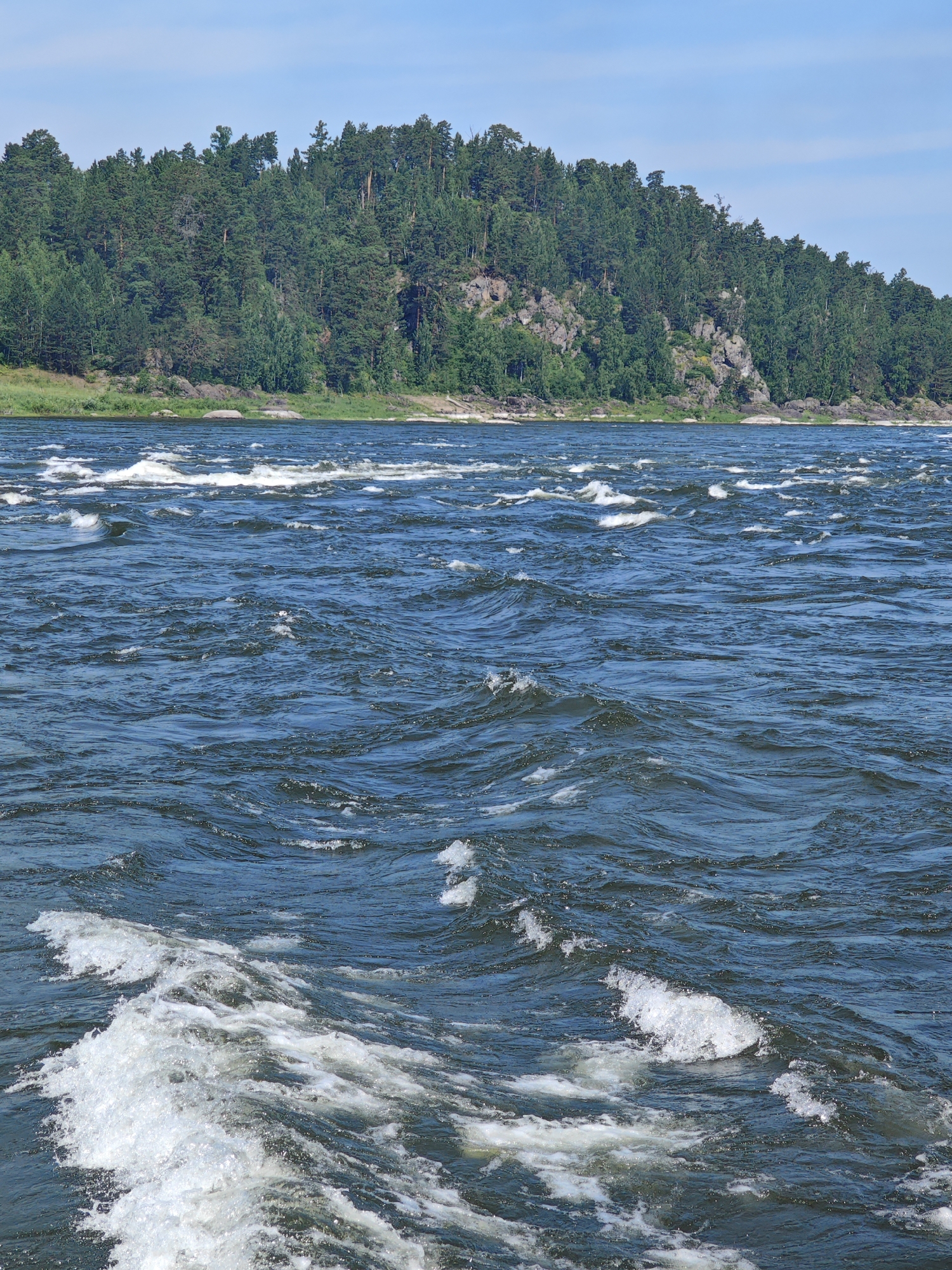 The image shows a wide, dark blue river with white-capped rapids in the foreground and middle ground. The river flows towards the viewer, with turbulent water creating frothy white foam. In the background, a densely forested hill rises from the riverbank, covered in evergreen trees and dotted with rocky outcrops. The sky above is a clear, pale blue. The scene appears to be a natural landscape, likely in a wilderness area. The city of Porog, Russia is known for its hydroelectric power station and surrounding natural beauty, and this image is consistent with the type of scenery found in that region.