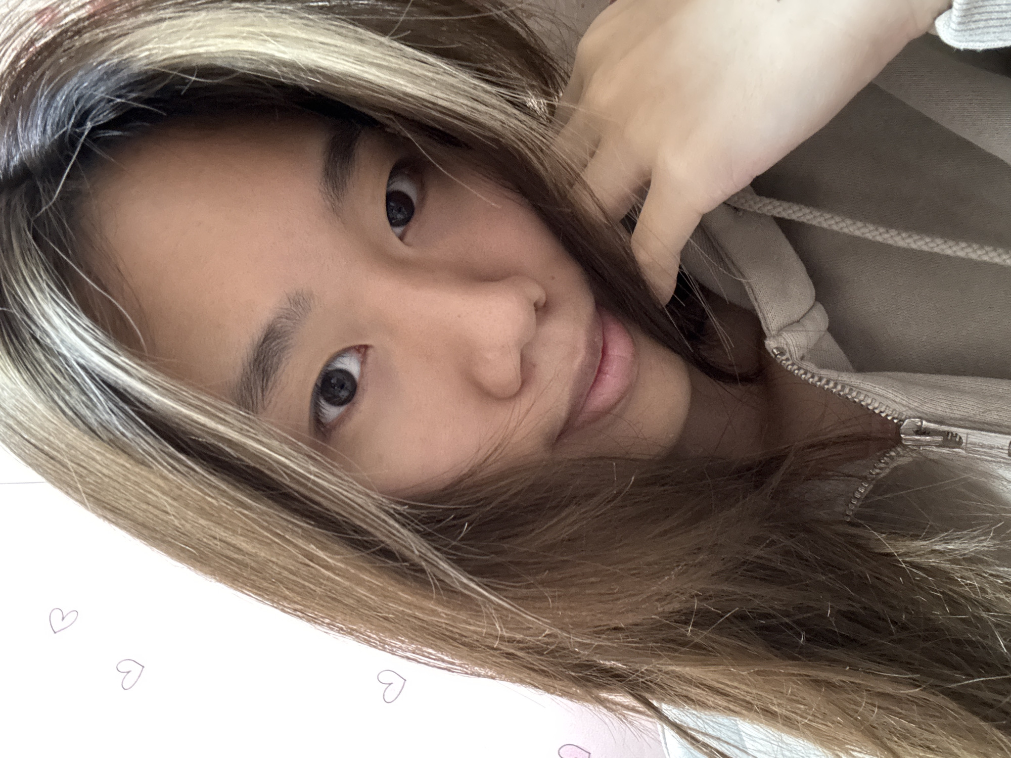 A young woman with brown and blonde hair is the central subject of the image. She appears to be wearing a beige hoodie. Her left hand is near the collar of the hoodie. The background includes white paper with faint heart drawings. There is no information present in the image to suggest the location is Shapingba District, China.