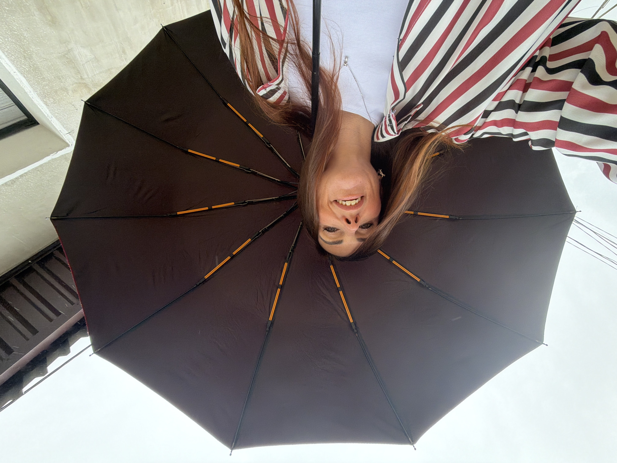 A woman with long, reddish-brown hair and dark eyes smiles widely, holding a large, open, dark-colored umbrella over her head. She is wearing a white t-shirt and a striped jacket with black, white, and red stripes. The image is taken from a low-angle perspective, looking up towards the umbrella and the sky. Above the umbrella, a portion of a building with a textured light-colored wall and dark window frames is visible, as well as what appears to be a wooden overhang or roof structure. The background is a bright, overcast sky, suggesting it might be a cloudy or rainy day.