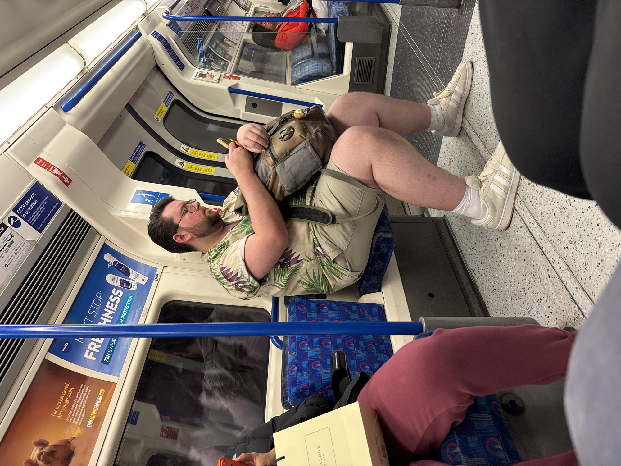 The image shows the interior of a London Underground train carriage. A man in a floral shirt and khaki shorts is seated, occupying two seats, with his legs crossed and a bag on his lap. He wears glasses and has a beard. Another person is partially visible in the foreground, wearing burgundy trousers and next to a "Jo Malone London" shopping bag. In the background, a seated passenger in an orange shirt can be seen. Advertisements are visible along the carriage wall, including one for Nivea deodorant with "NEXT STOP: FRESHNESS" and another highlighting the gassing of pigs, with the website ENDFACTORYFARMING.COM. Signs related to CCTV cameras are also visible. The blue seat upholstery and the blue handrail are characteristic of the London Underground.