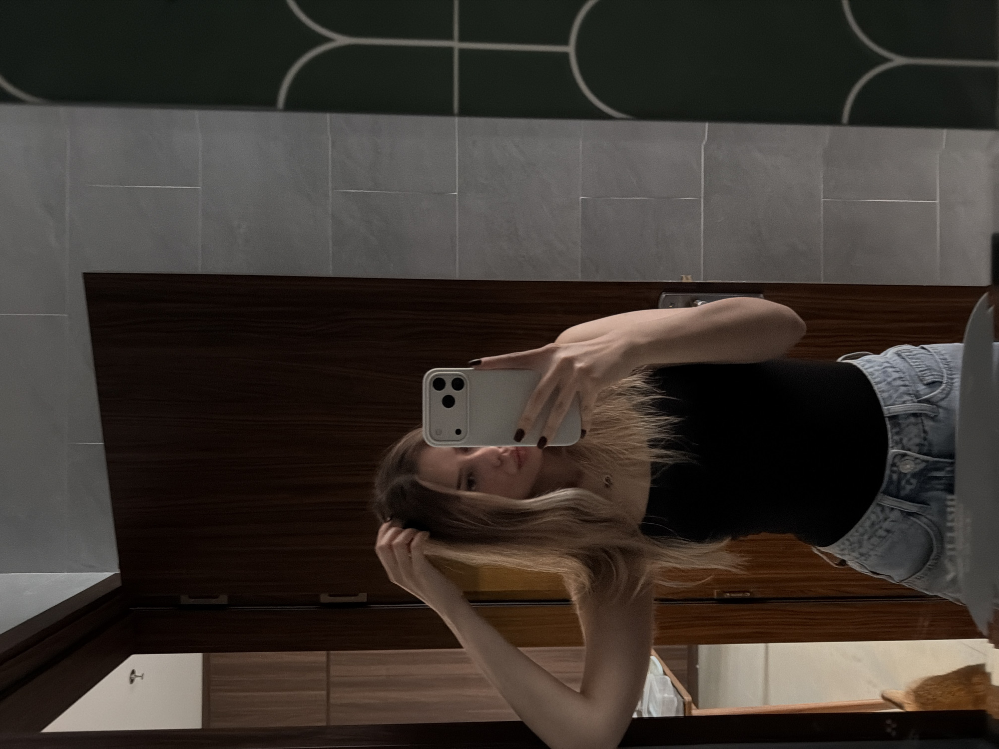 An unidentified woman is standing in a bathroom and taking a mirror selfie. She is wearing a black strapless top and light blue jeans. Her blonde hair is partially pulled up with her left hand. She is holding a light-colored smartphone in her right hand, with her index finger pressing the shutter button. The bathroom has a dark wooden door on the left, grey tiled walls, and a dark green wall with a white arched pattern on the right. The scene is indoors, likely in a hotel or residential setting. There is no visible indication that this location is in Đà Nẵng, Vietnam.