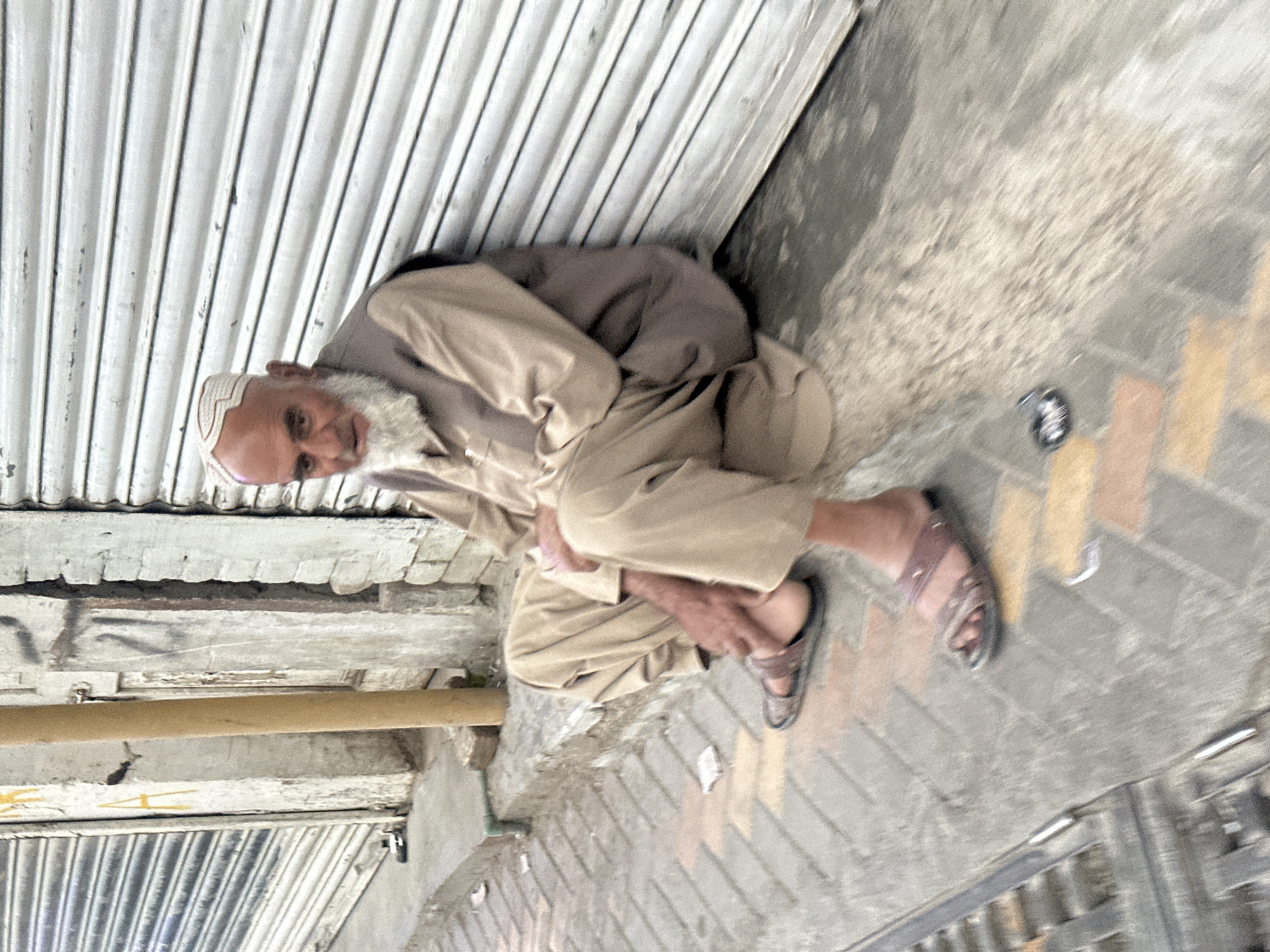 An elderly man in Quetta City Tehsil, Pakistan, crouches against a closed corrugated metal storefront. He wears a beige shalwar kameez, a white cap, and brown sandals. He is fair-skinned with a white beard. The street is paved with bricks and concrete. A metal bar runs along the wall above him.