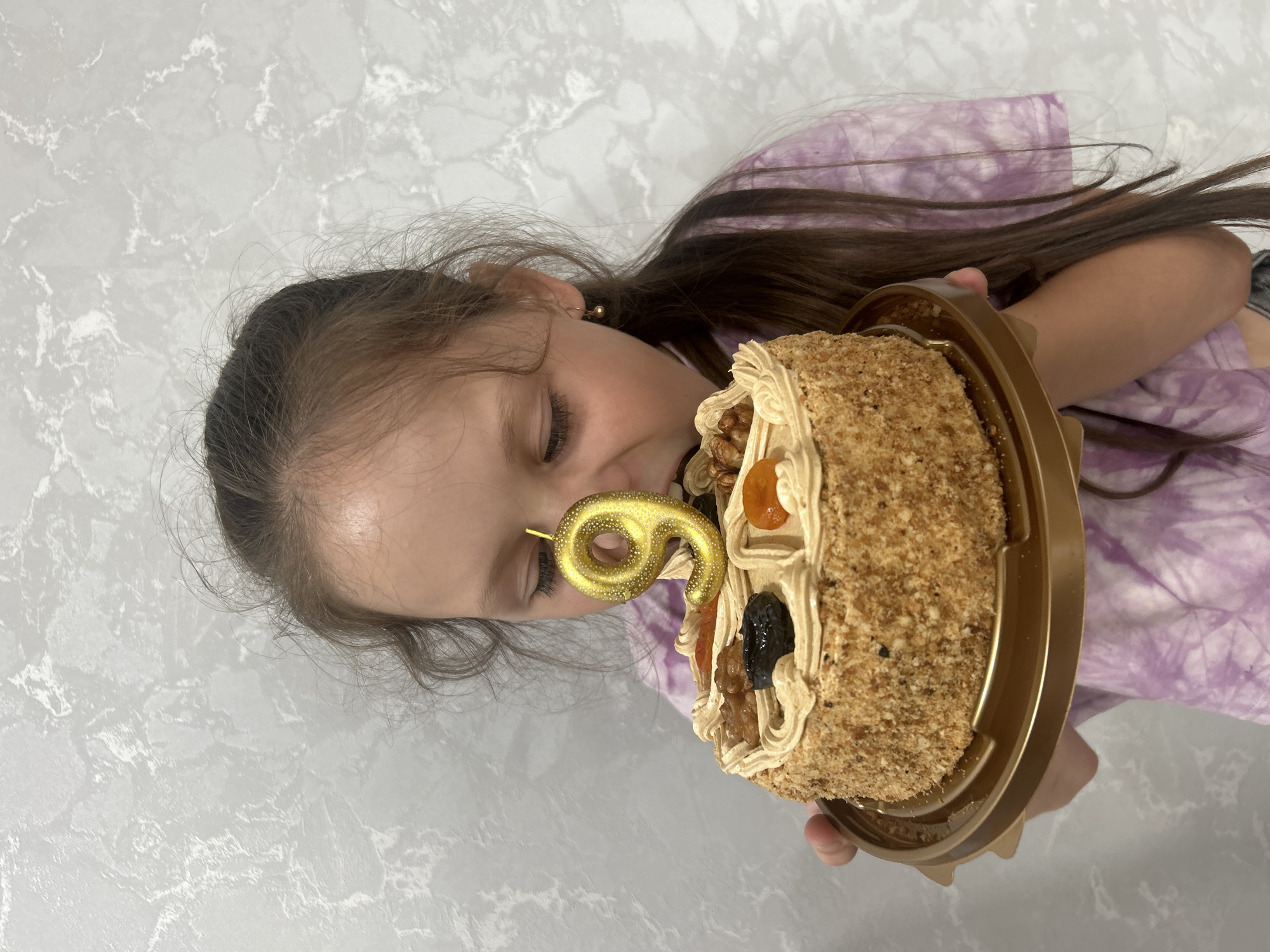 A young girl is holding a cake and about to eat it. The cake is decorated with frosting, nuts, and dried fruit, and has a gold candle in the shape of the number 9. The girl is wearing a purple tie-dye t-shirt. The background is a textured gray wall. The image appears to have been taken indoors.