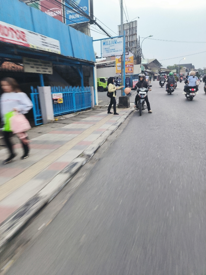 The image shows a street in Cileunyi, Indonesia (latitude: -6.933, longitude: 107.726). There is a woman wearing a black jacket and a white hijab standing on the sidewalk, holding an orange object in her hand. Behind her, there is a man wearing a blue helmet riding a motorcycle towards the left. There are other motorcycles on the street and several people walking on the sidewalk. The sidewalk is tiled, and the street is paved with asphalt. There is a building with a blue roof and a blue fence on the left side of the image. The building has a sign that says "William" in white letters on a red background. The sky is cloudy. The time of day is daytime. The weather is overcast.