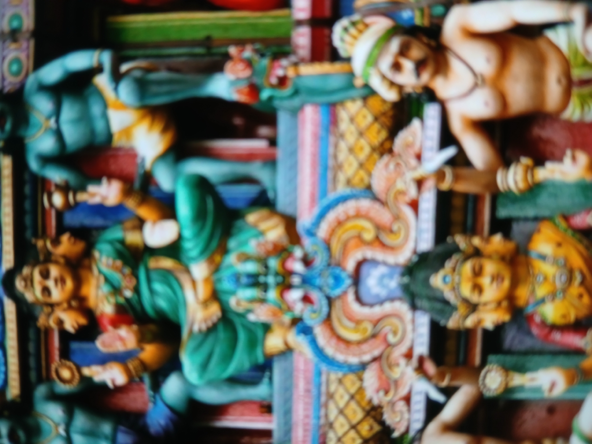 The image displays a section of a vibrant Hindu temple wall, likely located in Bengaluru, India. The scene features various sculpted figures of deities and characters from Hindu mythology. The figures are painted in bright colors like blue, green, yellow, and orange. They are adorned with jewelry and intricate detailing. The temple wall is decorated with patterns and designs. The general atmosphere indicates a place of religious significance and artistic expression. The figures are positioned in a stacked arrangement, with deities facing different directions.
