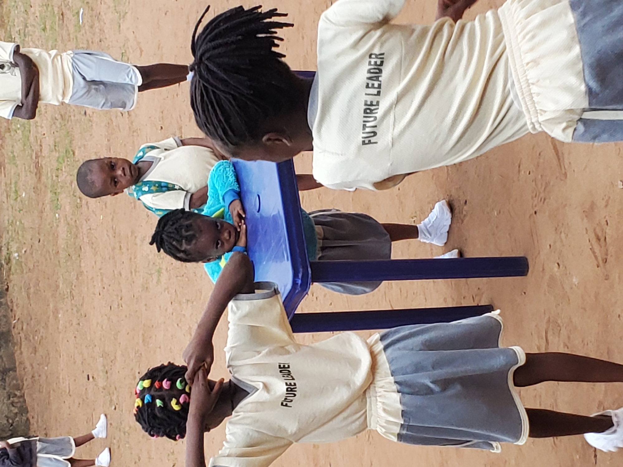 The image shows several children in Ukwu-Abwa, Nigeria, on what appears to be a dirt or sand-covered ground. All are wearing similar uniforms consisting of tan shirts with the words "FUTURE LEADER" printed on the back. Some wear shorts, while others wear skirts, both of a grey color with white trim. At least one child is leaning on or sitting at a blue plastic table. Others are standing around the table. The scene appears to be outdoors, possibly a schoolyard or recreational area.
