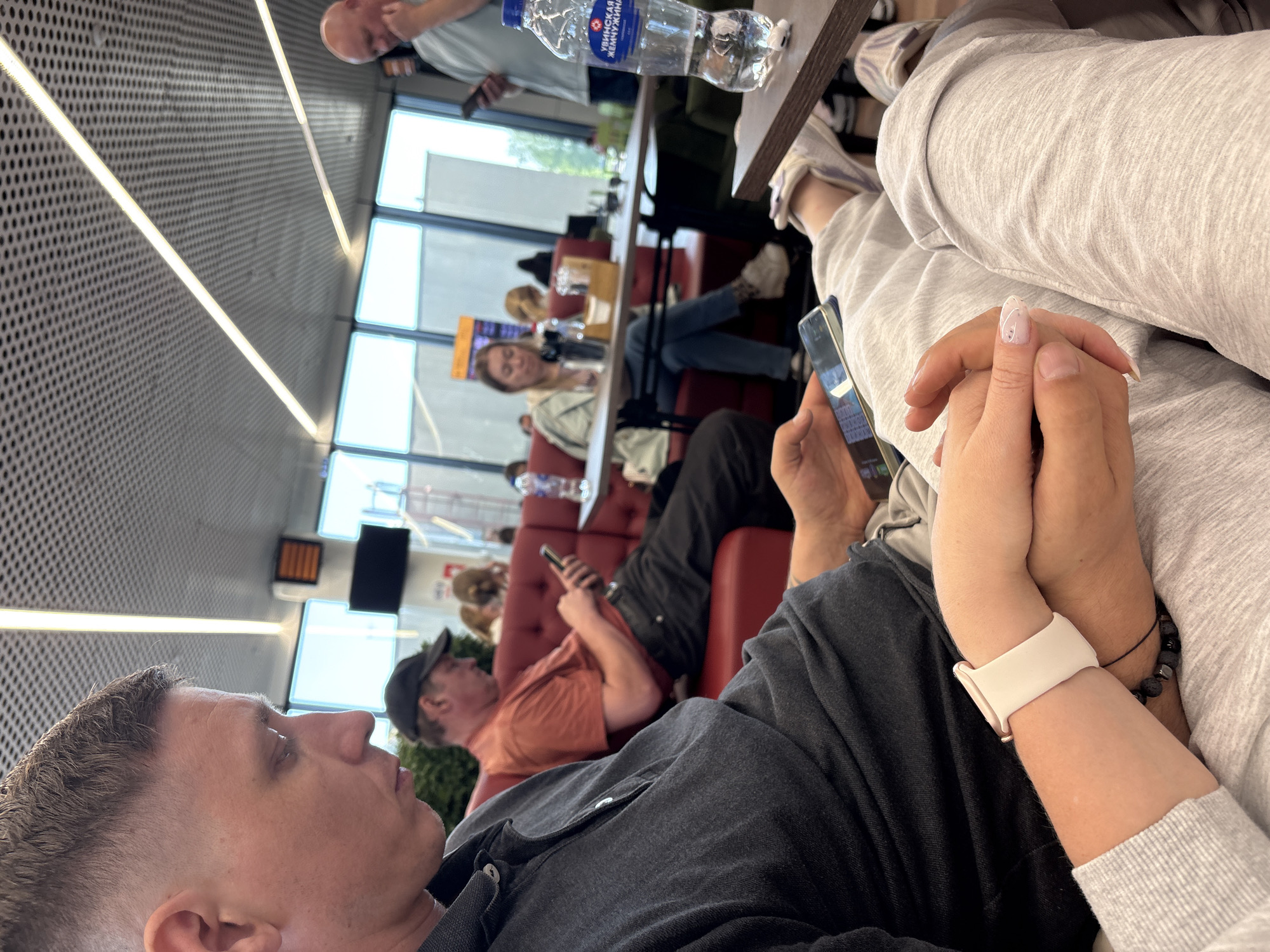 The image captures a scene in a waiting area, possibly at an airport or train station, in Новое Мартьяново, Russia. Several individuals are present, seated in red upholstered chairs, seemingly relaxing or waiting. A man in the foreground is partially visible, with a close-up of his face showing a short haircut. Adjacent to him, a woman is holding a smartphone in one hand, and her other hand is held by someone else. She is wearing a white smartwatch and has a bracelet with black beads on her wrist. In the background, other people are also visible, some looking at their phones. There are tables present, one with a bottle of water on it. The environment suggests a public space with rows of chairs and windows providing natural light.