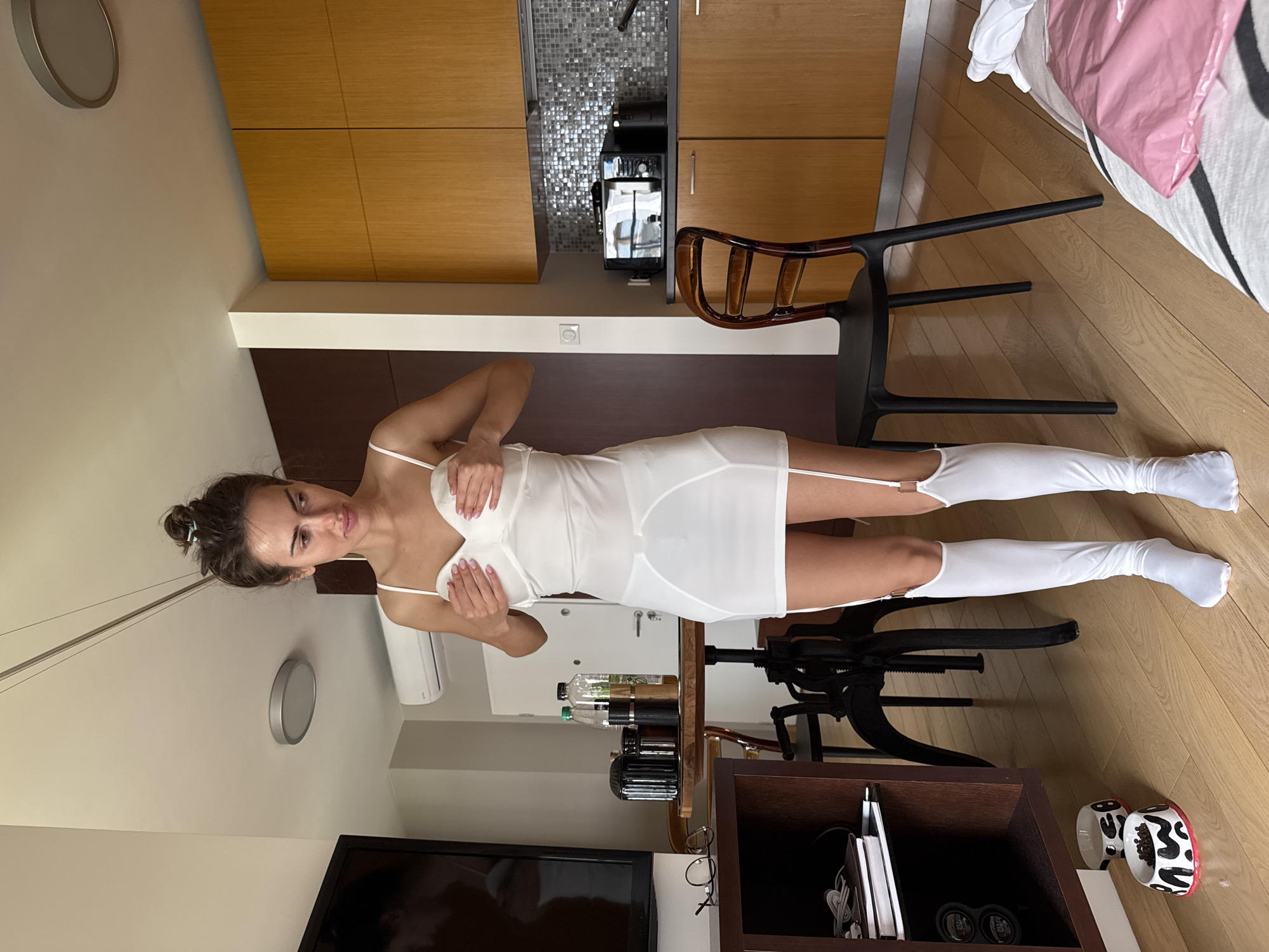 A woman is posing indoors in a white dress and white thigh-high stockings. She stands near a dark wood table with several bottles and a coffee maker on it. A TV is in the lower left corner of the frame, and a round wall clock is visible on the wall behind her. The background includes a kitchen area with wooden cabinets and a patterned backsplash, and a chair is positioned nearby. The flooring appears to be wooden.