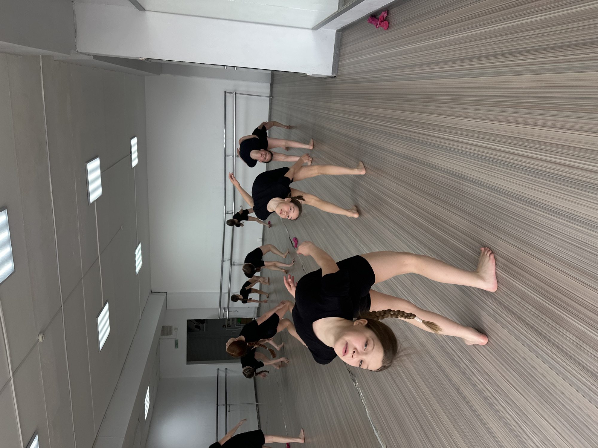 The image depicts a dance studio in what can be reasonably assumed to be Votkinsk, Russia, given that information was provided. Several young girls are practicing dance poses. They are wearing black attire. The studio has a mirrored wall along one side with a barre, and the floor is made of light-colored wood. Artificial lighting is provided by ceiling-mounted fluorescent fixtures.