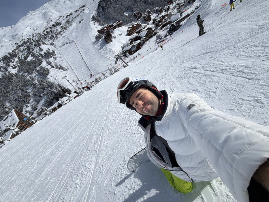 The image presents a first-person, tilted view of an individual on a snow-covered ski slope within the Les Allues commune in France. The central subject, an adult male, is positioned in the foreground, captured in a selfie. He wears a white quilted ski jacket over grey and black layers, bright lime green ski trousers, and a black and white 'Atomic' branded ski helmet with goggles resting on top. A snowboard is partially visible to his left. He has his eyes closed and a slight smile, with his right arm extended to hold the camera. The expansive, well-groomed slope exhibits numerous ski and snowboard tracks. Further down the slope, multiple individuals are visible, including a skier in dark clothing and another wearing yellow pants. The background displays a ski resort village composed of numerous snow-covered, chalet-style buildings interspersed with dense evergreen forest. Ski lifts and a large cleared snow area are discernible within the village complex. Distant snow-covered mountains frame the scene under a clear, bright blue sky.