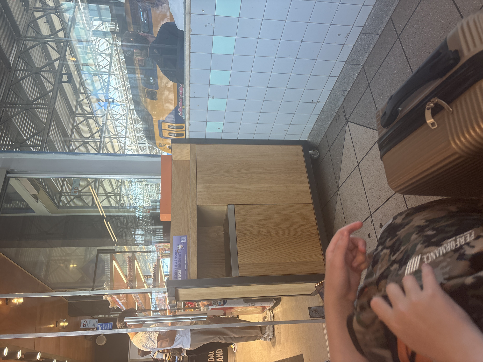 The image captures an indoor scene, likely a waiting area or a shop within a transportation hub, possibly Eindhoven Centraal station. A yellow and blue train is visible through a large glass window, indicating a train station environment. In the foreground, a person's hands are visible, resting on a camouflage-patterned backpack. To the right, a gold suitcase is placed on the tiled floor. An orange object, possibly a box, sits atop a wooden and metal counter unit, which is part of a display or service area. Behind the counter, shelves stocked with various items are visible through a glass partition. A security camera is mounted on a blue sign to the left. The overall lighting suggests an interior space with natural light filtering in from the exterior.