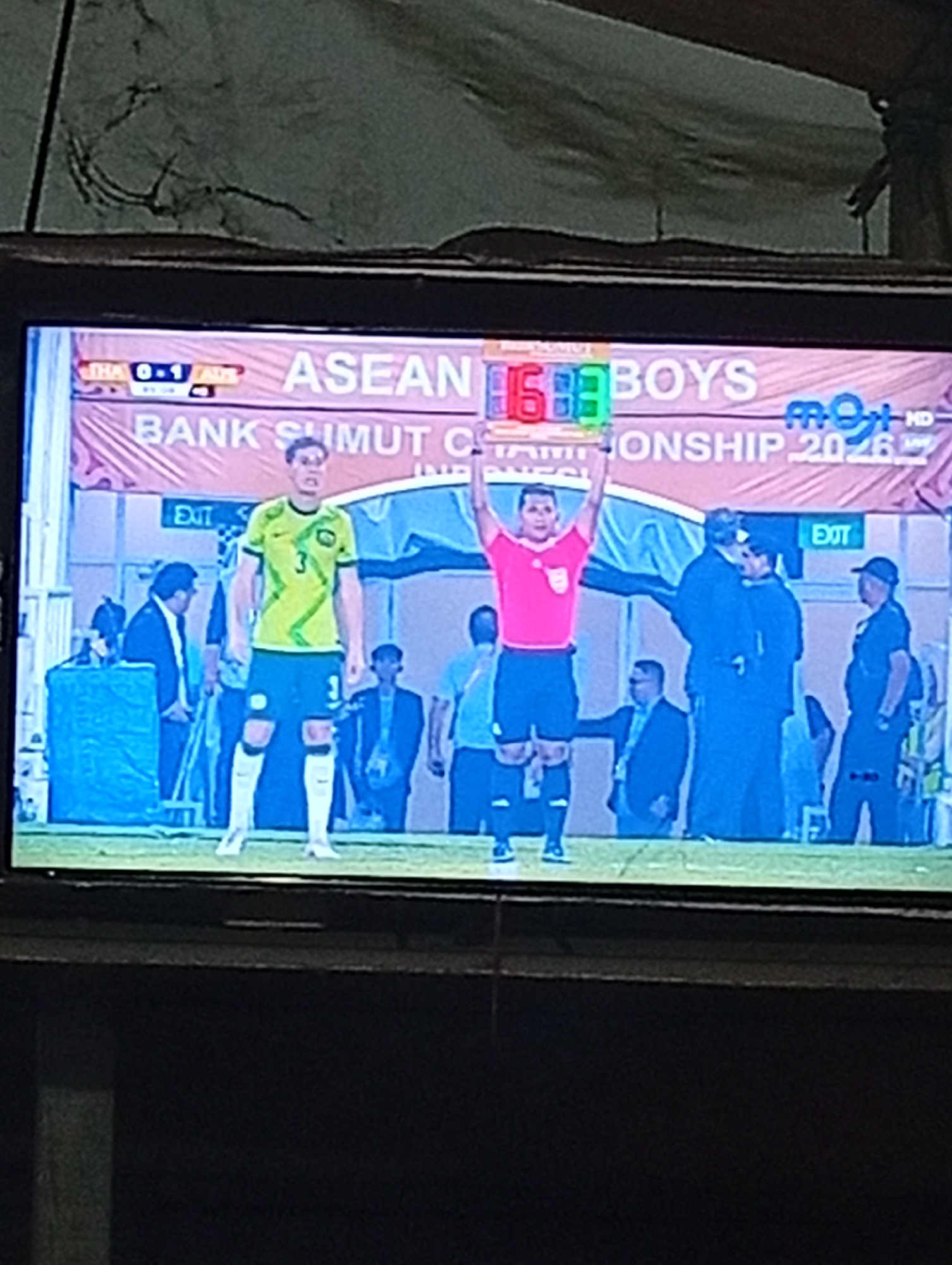 The image captures a moment from a televised football match, likely broadcast from Lhokseumawe, Indonesia, as indicated by the "BANK SUMUT" sponsor visible on the banner behind the players. The screen shows a football player in a lime green and yellow jersey with the number 3 on his shorts and jersey, standing on a green field. To his right, a referee in a pink shirt and black shorts is holding up a substitution board displaying the numbers 6 and 3, indicating a player substitution. Behind them, several officials and spectators are visible, some in suits and others in casual attire. The overall scene is a typical sporting event broadcast, with the score of "THA 0-1 ADS" displayed on the top left of the screen.