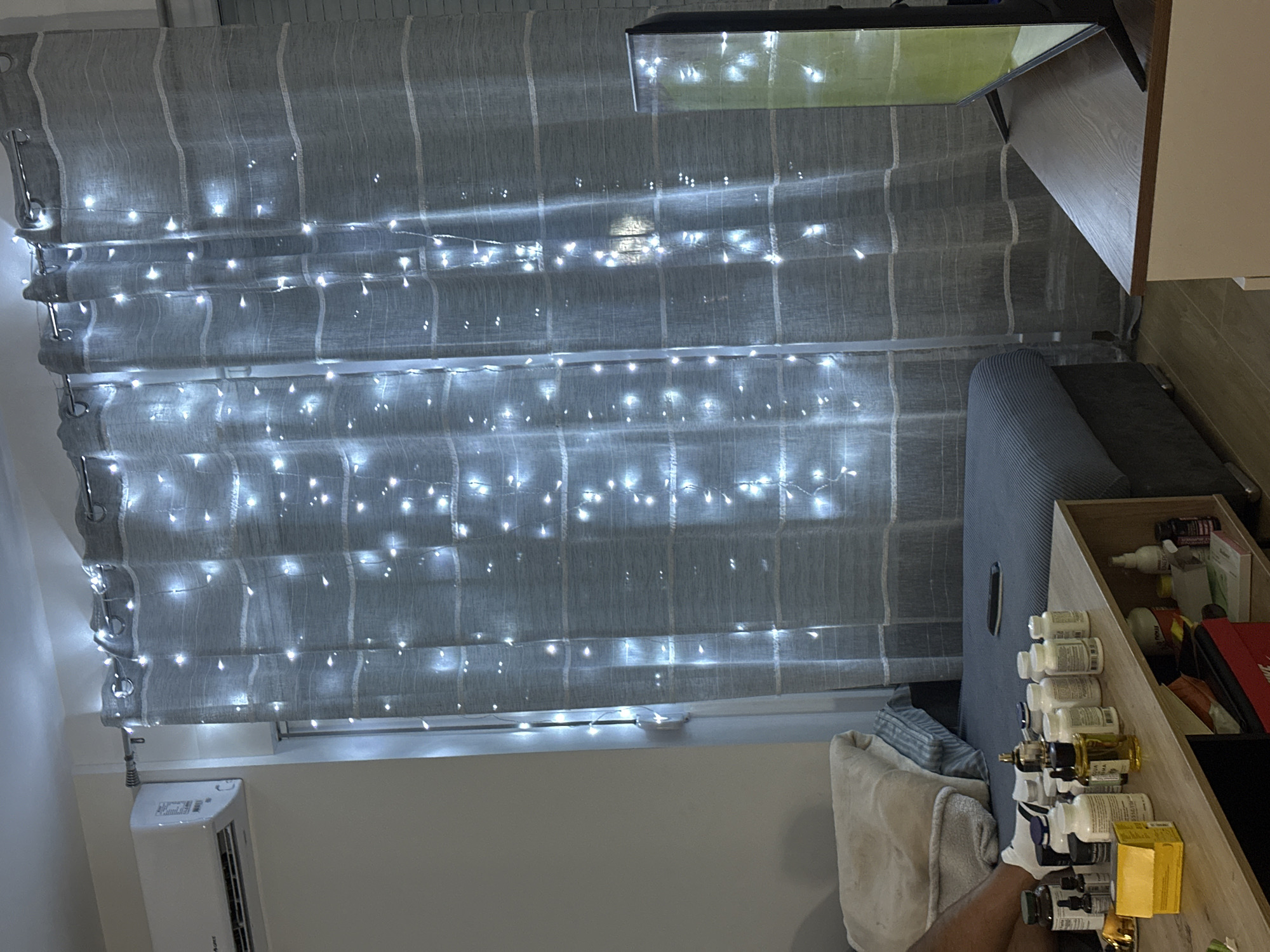 In Donji Pobori, Montenegro, an interior scene depicts a living area with a sofa and a coffee table. The room is illuminated by a string of white fairy lights draped across grey curtains, which are hanging from a curtain rod. To the left of the curtains, an air conditioning unit is mounted on a light-colored wall. On the coffee table, an assortment of pill bottles and other containers are arranged. A television screen is visible to the right, set on a wooden cabinet. The sofa is covered with a grey ribbed fabric, and a folded blanket rests on its left side. A person's legs and feet, wearing white socks, are visible on the left edge of the frame, resting on the sofa.
