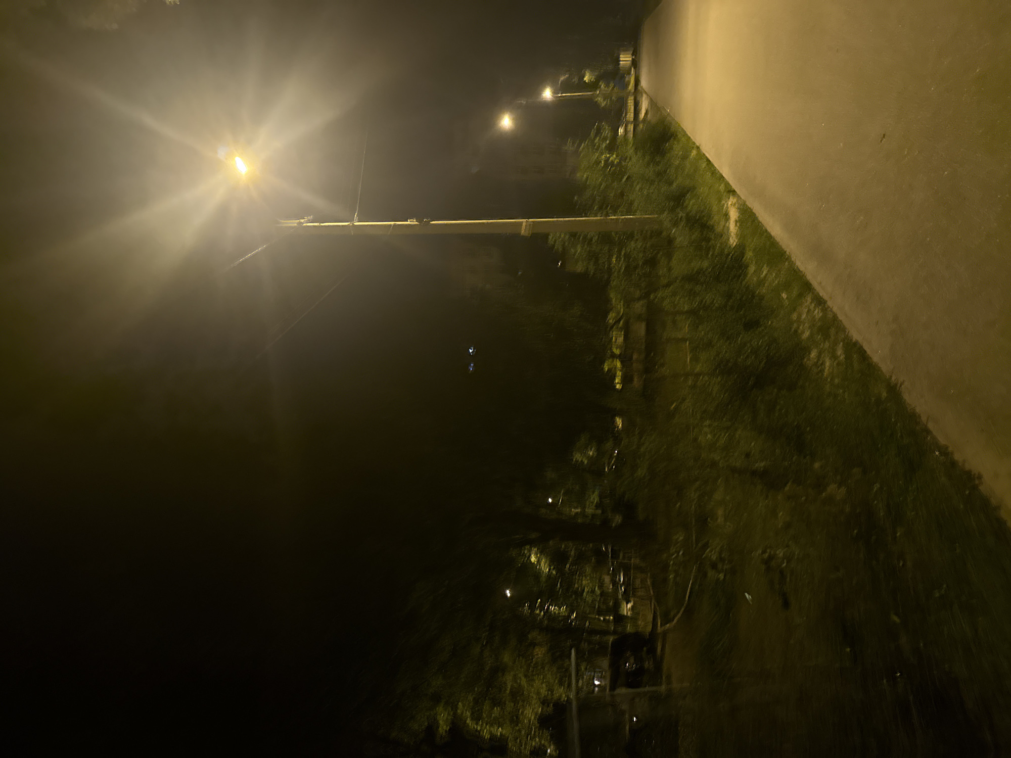 The image depicts a nighttime scene of a paved walkway adjacent to a grassy area with trees. A tall lamppost with a bright light illuminating the surrounding area is visible on the left side of the walkway. Further down the walkway, another lamppost is visible, also illuminated. The environment appears to be an urban park or residential area. The sky is dark, indicating it is nighttime. The general location is the city of Dnipro, Ukraine.