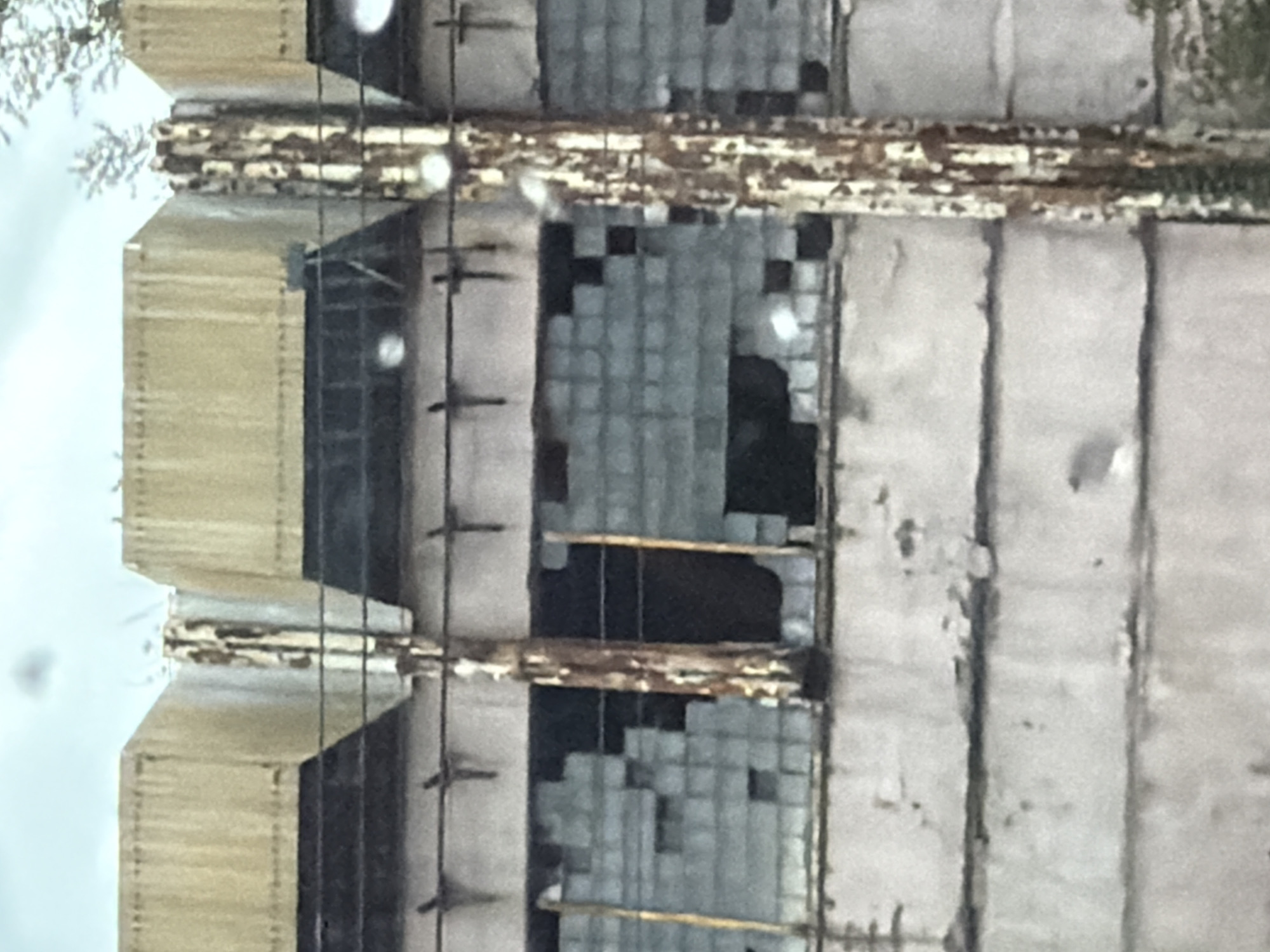 The image shows a weathered, industrial building facade in Kyiv, Ukraine. The building appears to be constructed of concrete blocks, with sections of large, glass-like panels, many of which are broken or missing, revealing dark interiors. Several large, rust-covered metal columns run vertically along the facade. Above these are what appear to be ventilation units or roof structures, with a textured, yellowish-brown surface, also showing signs of wear and tear. The sky is overcast. The overall impression is one of neglect and disuse.