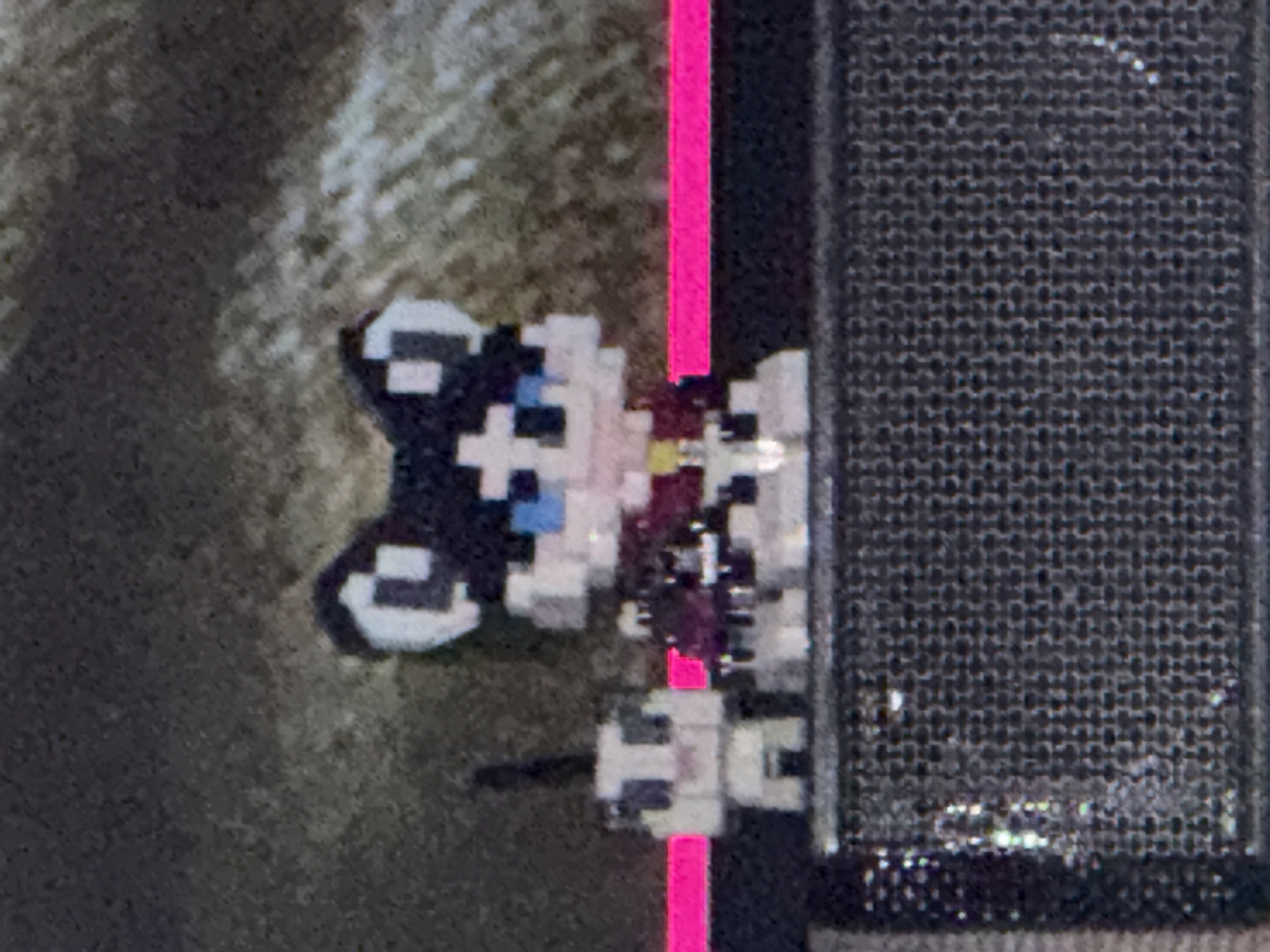 A pixel art representation of a cat, predominantly white with black markings around its ears and a blue eye, is positioned adjacent to a black, textured rectangular object. A vertical pink line bisects the space between the pixel art cat and the rectangular object. The background appears blurry and textured, possibly concrete or a similar rough surface. The location is unknown.