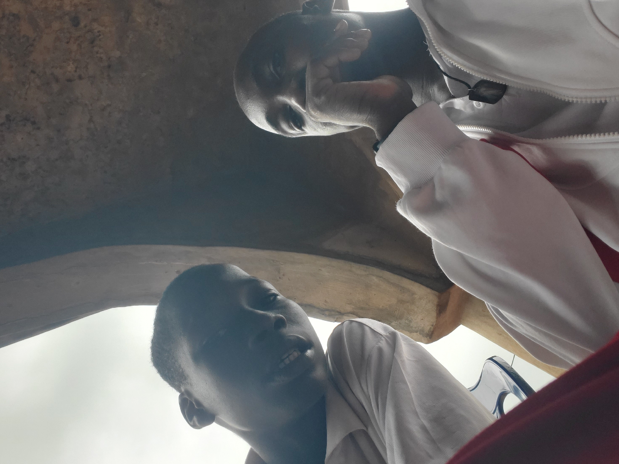 This is a low-angle, close-up selfie taken from a low perspective, showing two young Black men against a backdrop that appears to be an outdoor, possibly covered area with a textured, earthy-toned ceiling or overhang. The lighting is diffused and appears to be daylight, creating soft shadows.
The man on the left is positioned closer to the camera and his face is mostly in shadow, but his mouth is slightly open, revealing teeth, and he has a faint smile. He is wearing a white garment, possibly a hoodie or jacket, and a bit of a red garment is visible in the lower left corner, possibly pants or a shirt. His left ear is visible.
The man on the right is slightly further back and his face is more clearly visible. He is resting his right hand on his chin and looking directly at the camera. He has a neutral expression. He is wearing a white zipped jacket over a white shirt, and a dark pendant necklace is visible on the shirt.
The environment suggests an urban or semi-urban setting in Nsugbe, Nigeria. The rough, textured ceiling could be part of a building's architecture, a market stall, or an outdoor recreational structure. The presence of a blue plastic chair is visible between the two men, suggesting a casual, perhaps public, gathering place. The overall impression is of a candid moment captured by the individuals themselves.