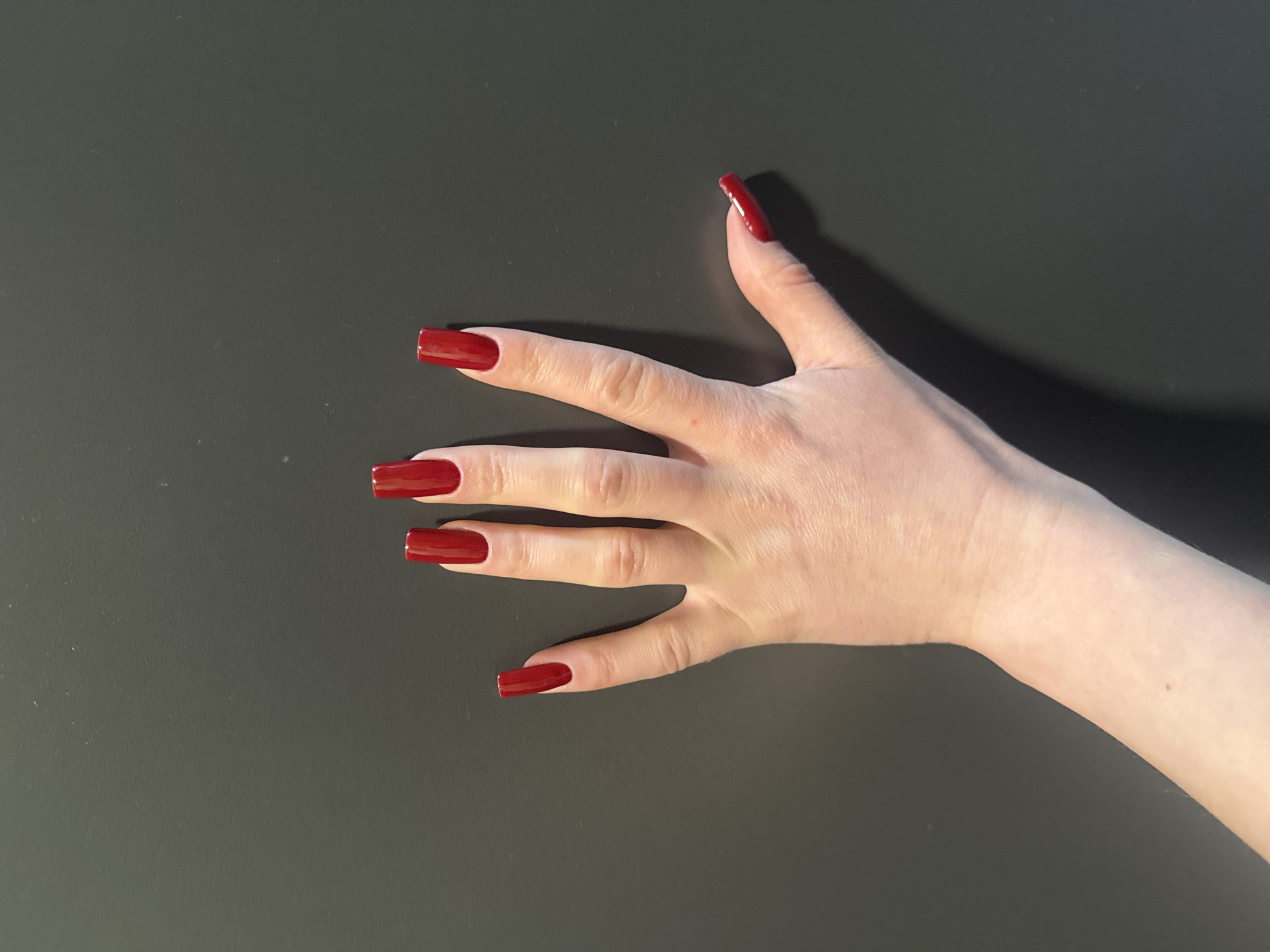 A fair-skinned left hand is presented against a plain, dark gray background. All five fingers are spread, with the palm facing downwards towards the unseen surface. Each fingernail is long, rectangular with slightly squared-off tips, and coated in a glossy, deep red nail polish. No jewelry or distinguishing marks are visible on the hand. The background is uniform in color and texture, showing minor surface imperfections. The lighting is even, casting subtle shadows to the right and beneath the hand. The specific location in the city of Unknown, Unknown, cannot be determined from the visual information provided; the scene appears to be an indoor, controlled environment.