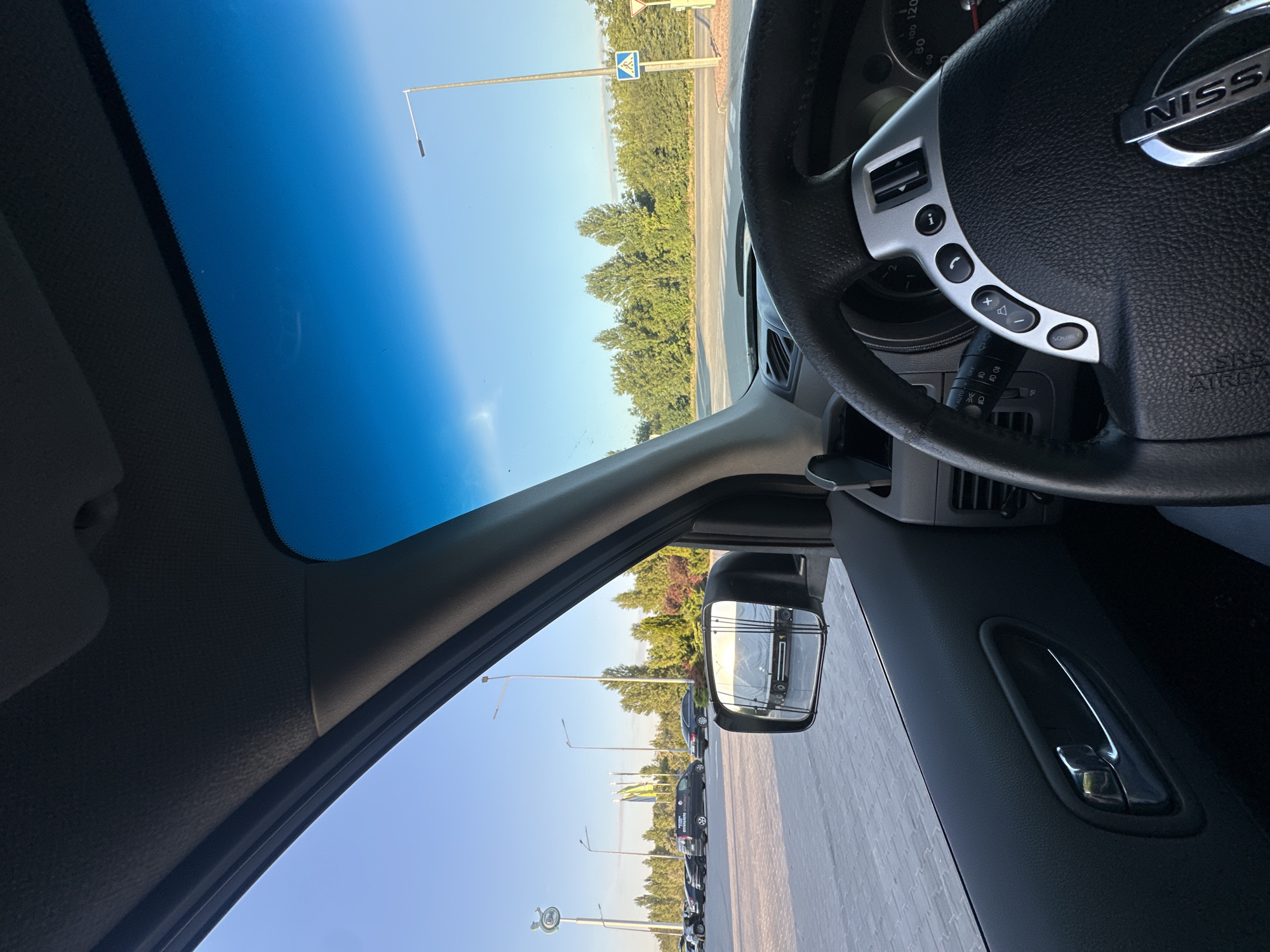 The image is taken from inside a car, looking forward and slightly to the left. The driver's side interior, including the steering wheel and door handle, is visible. Outside, a parking lot is seen with several cars and a truck in the side mirror. The background features a line of trees and a clear blue sky. The location is identified as Kamianets-Podilskyi, Ukraine.