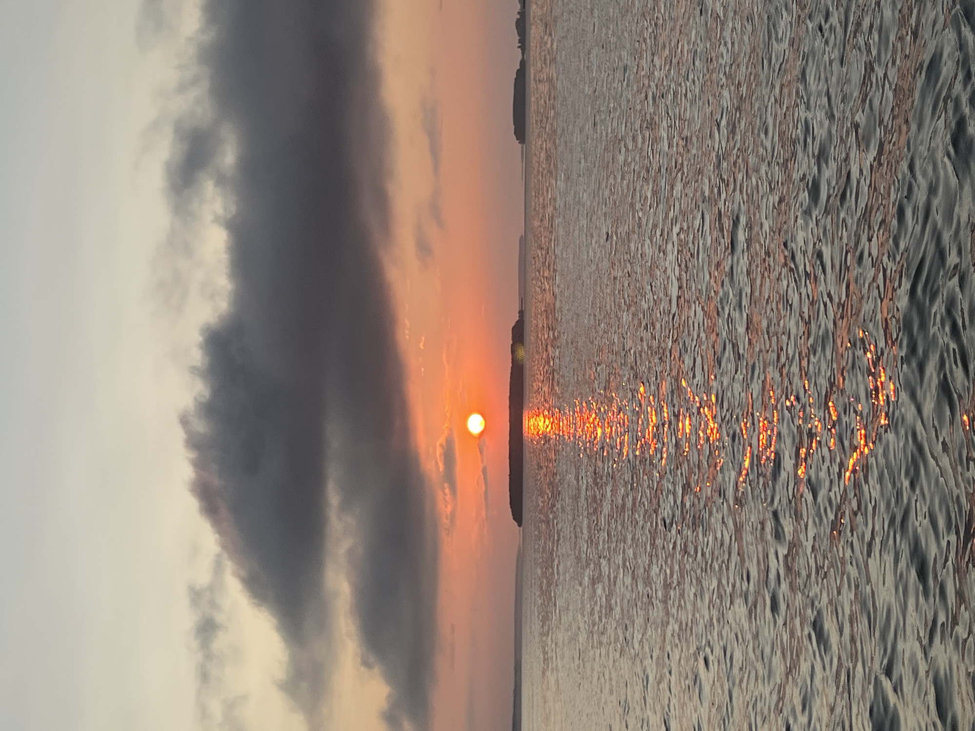 The image depicts a tranquil sunset over a body of water, likely a lake or a bay. The sun, a bright orange orb, is low on the horizon, casting a vibrant reflection that shimmers across the rippling surface of the water. Dark, heavy clouds dominate the upper portion of the sky, tinged with hues of orange and pink from the fading sunlight. In the distance, silhouetted islands or peninsulas are visible against the colorful sky, adding depth to the scene. The overall atmosphere is one of serene natural beauty. The location is identified as Novopichugovo, Russia.