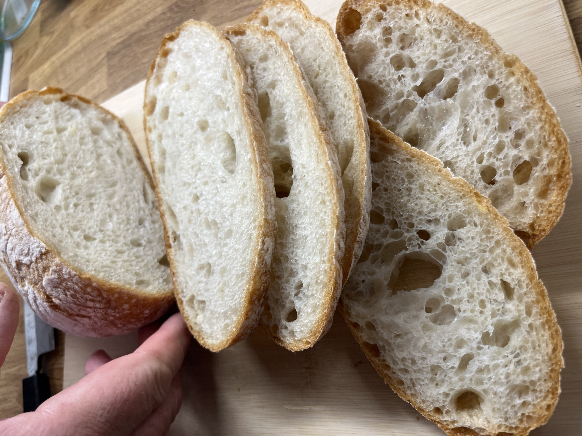 In a close-up, eye-level shot, a freshly baked loaf of rustic bread sits on a wooden cutting board. The loaf is sliced, revealing a porous, airy crumb and a golden-brown crust dusted with flour. Several slices lie stacked on the board, their textures and colors highlighted by natural light. A hand, partially visible in the upper left, reaches towards the loaf, suggesting it has just been cut or is about to be served. The background shows a wooden surface, possibly a kitchen counter or table. The scene is devoid of any discernible urban elements, indicating it is likely indoors, perhaps in a home kitchen or bakery. There are no visible landmarks or context to place this image specifically within Helsinki, Finland.