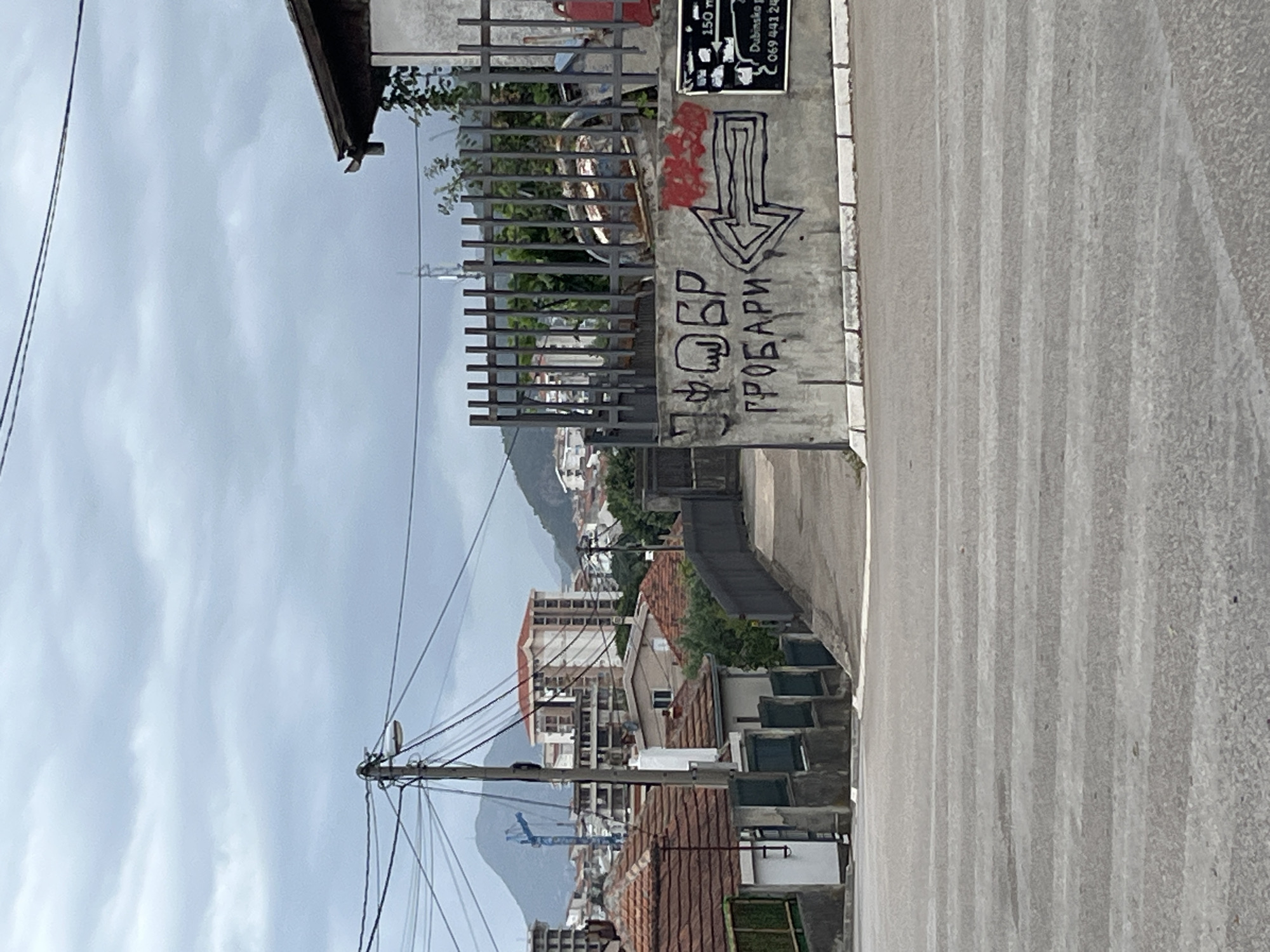 The image shows a street view with buildings and a cloudy sky. In the foreground, there is a street with white lines. On the right side of the image, a concrete wall with graffiti is visible. The graffiti includes Cyrillic text and an arrow pointing to the left. There is also a sign with some text and numbers. Behind the wall, a metal fence is present, and behind that, there are trees and more buildings. The background shows a cityscape with residential and apartment buildings, as well as a large mountain. There is a construction crane visible in the distance. The overall impression is that of an urban environment on a cloudy day.