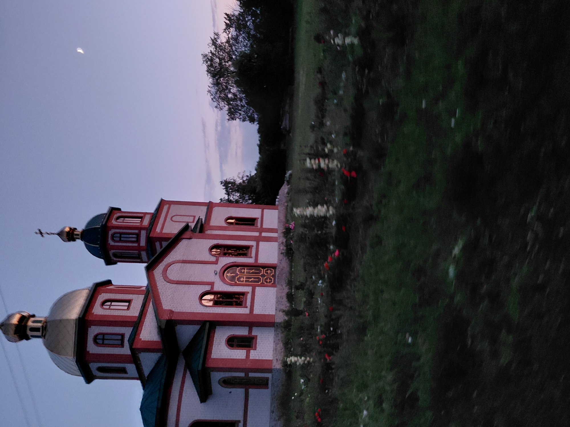 The image depicts an Orthodox church in Ustyvytsia, Ukraine, at dusk. The church is constructed with white brick and red trim, featuring a prominent golden dome topped with a cross. A smaller dome with a cross is visible on a side tower. The windows of the church are illuminated, reflecting the twilight sky. In front of the church, there is a garden with scattered flowers. The background consists of dark trees and a pale twilight sky with a sliver of the moon visible. The foreground shows dark, out-of-focus vegetation. No people are visible in the scene.