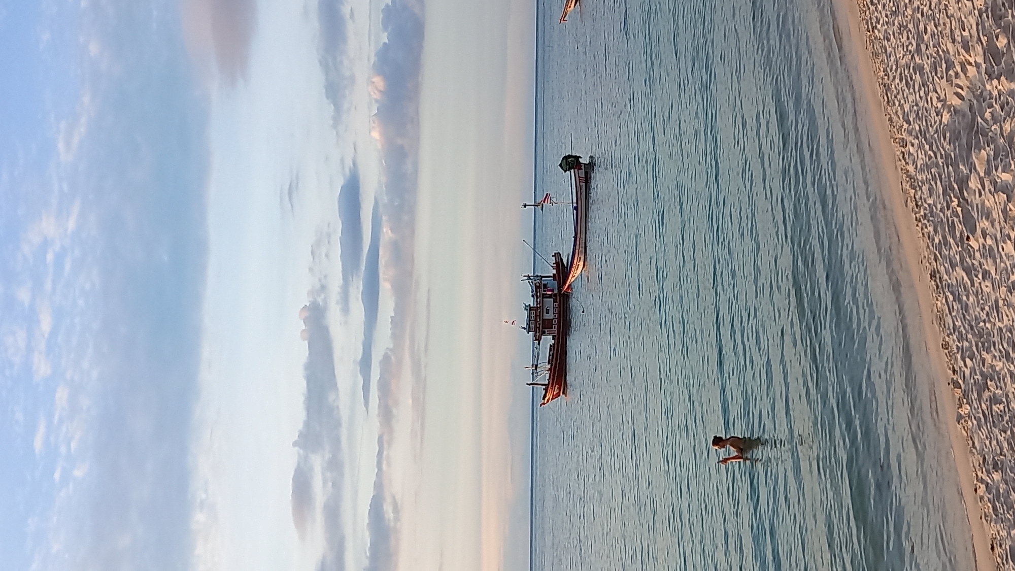 This media file shows a beach scene at Koh Phangan, Thailand, likely during sunset. The image is oriented vertically. On the right side of the frame is a sandy beach with footprints. The rest of the image is filled with the ocean, which is a calm, light blue with ripples.
In the water, a person is partially submerged, appearing to be wading or swimming. Further out in the ocean, there are two longtail boats. The first boat is closer to the shore, and the second is further out, appearing to be behind the first. Both boats have a traditional Thai design, with a long, narrow hull and an elevated platform at the stern.
The sky is a soft blend of blues and grays, with scattered clouds that are illuminated by what appears to be the setting sun, casting a warm glow on the scene. The overall mood is peaceful and serene, suggesting a quiet evening by the sea. There is no visible text in the image.