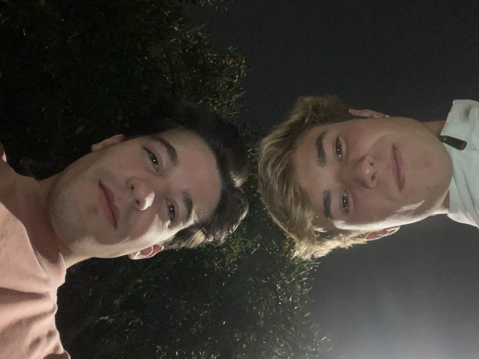 The inverted image captures two young adult males in a close-up, low-angle selfie at night in Aldoar, Foz do Douro e Nevogilde, Portugal. The upper subject (appearing at the top if rotated 180 degrees) has dark brown, medium-length hair and wears a light pink t-shirt. He faces the camera with a subtle smile. Below him (appearing at the bottom if rotated) is a male with light brown, short hair, wearing a light-colored collared jacket with a zipper. He exhibits a subtle smile and a small hoop earring in his visible right ear. The background is dark, consistent with nighttime, and includes blurred dark green foliage.