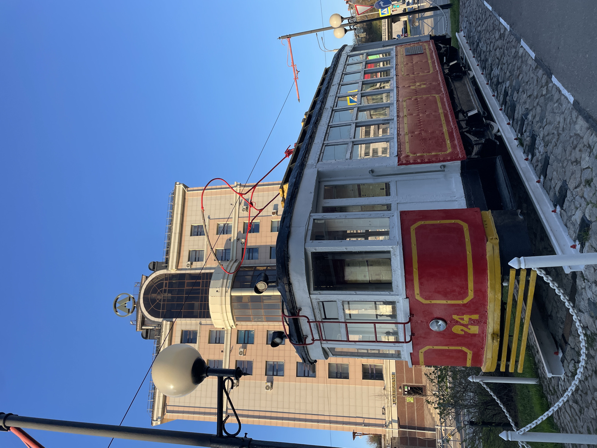A stationary, vintage, red and white tram, numbered 24 with yellow accents, is on display. The tram is parked on a cobblestone path and secured by a white fence with a chain. A modern multi-story building with numerous windows and a circular logo on top is in the background. An ornate street lamp stands near the front of the tram. The sky is clear blue. The location is likely Kazan, Russia, as indicated by the street name "OCTOROBAH" on the building.
