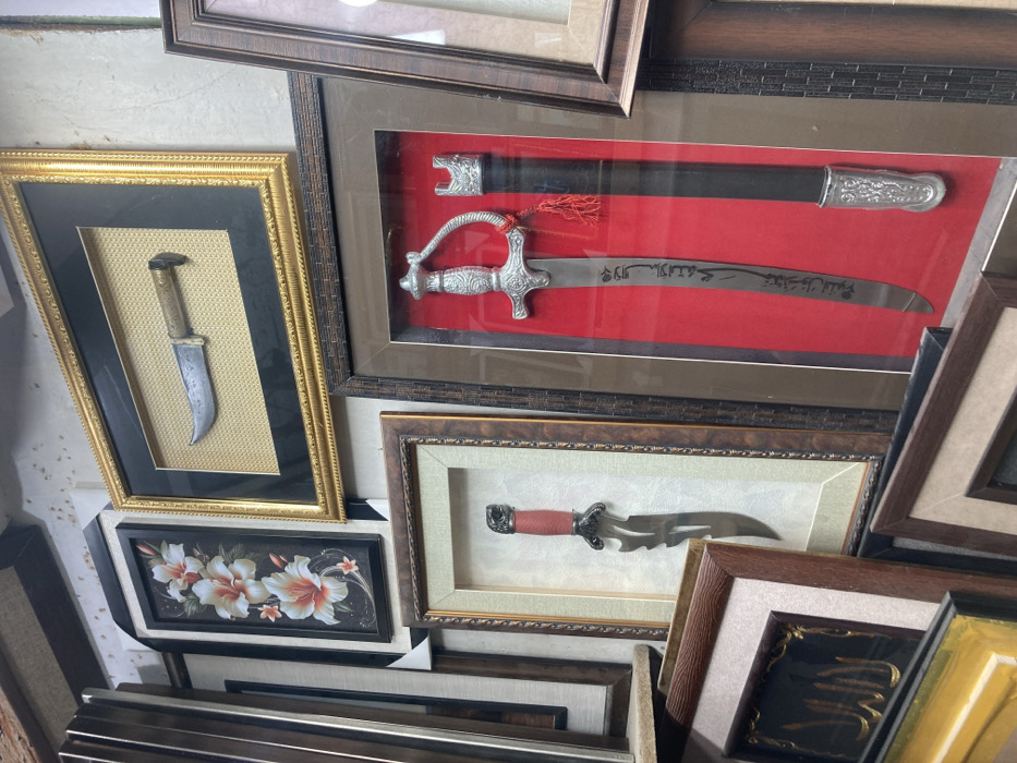 The image presents a detailed view of multiple framed decorative objects displayed in what appears to be an indoor commercial setting, likely a shop or gallery, located in Quetta City Tehsil, Pakistan. No individuals are visible.
The central focus is a cluster of framed items, prominently featuring edged weapons. In the upper right quadrant, a horizontal, ornate gold-colored frame encases a curved-blade knife with a light-colored handle, displayed against a light-brown textured background. Below and to the right of this, a large, dark-brown ornate vertical frame showcases a decorative sword with an elaborate silver-colored hilt and Arabic calligraphy engraved on its blade, accompanied by a black scabbard featuring matching silver-colored fittings and a red tassel, all set against a vibrant red velvet-like backing. To the left of the sword display, another dark-brown ornate vertical frame holds a dagger with a red handle and an intricately shaped, possibly serrated, blade, presented on a light-colored textured background.
In the upper left quadrant, a vertical rectangular frame with a black interior border and white exterior border displays artwork depicting three white and orange lilies with green stems on a dark background. Various other frames, some with dark wooden appearances, are partially visible, stacked and leaning against each other in the foreground and along the left edge of the image. One such partially visible frame in the lower left corner contains golden Arabic calligraphy on a dark surface. The items are arranged densely, suggesting a retail environment for decorative arts or traditional weaponry.