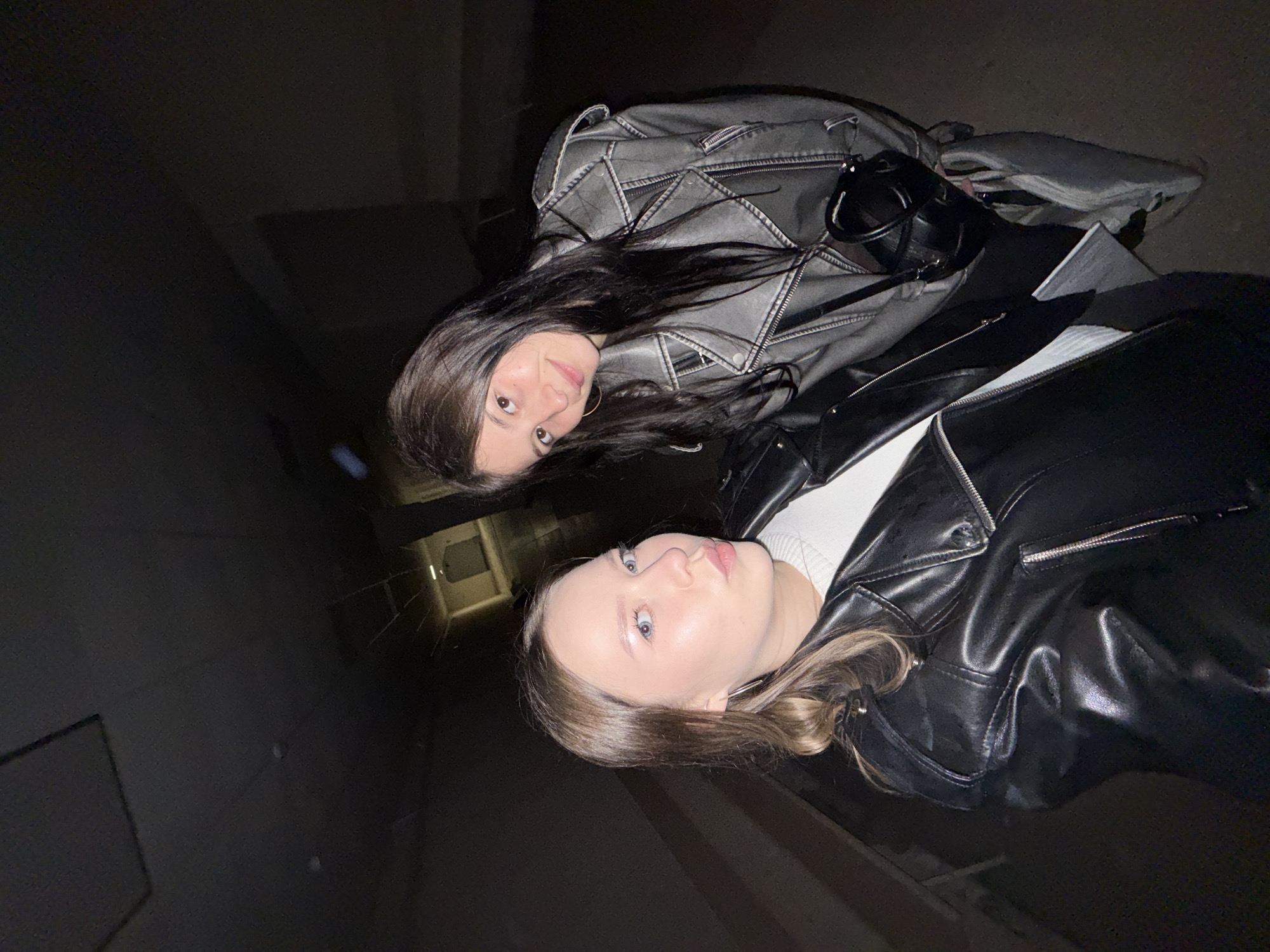 Two young women are standing in a dimly lit, narrow corridor or hallway, possibly an underpass or industrial building. The woman on the left, with blonde hair, is looking at the camera with her lips puckered and wearing a black leather jacket and a white top. The woman on the right, with dark hair, is smiling and looking slightly off-camera, wearing a grey leather jacket and carrying a black bag. The background is dark and indistinct, with some architectural features visible in the distance. The overall lighting suggests it is nighttime or the area is poorly illuminated. The scene is captured in Chaikovsky, Russia.