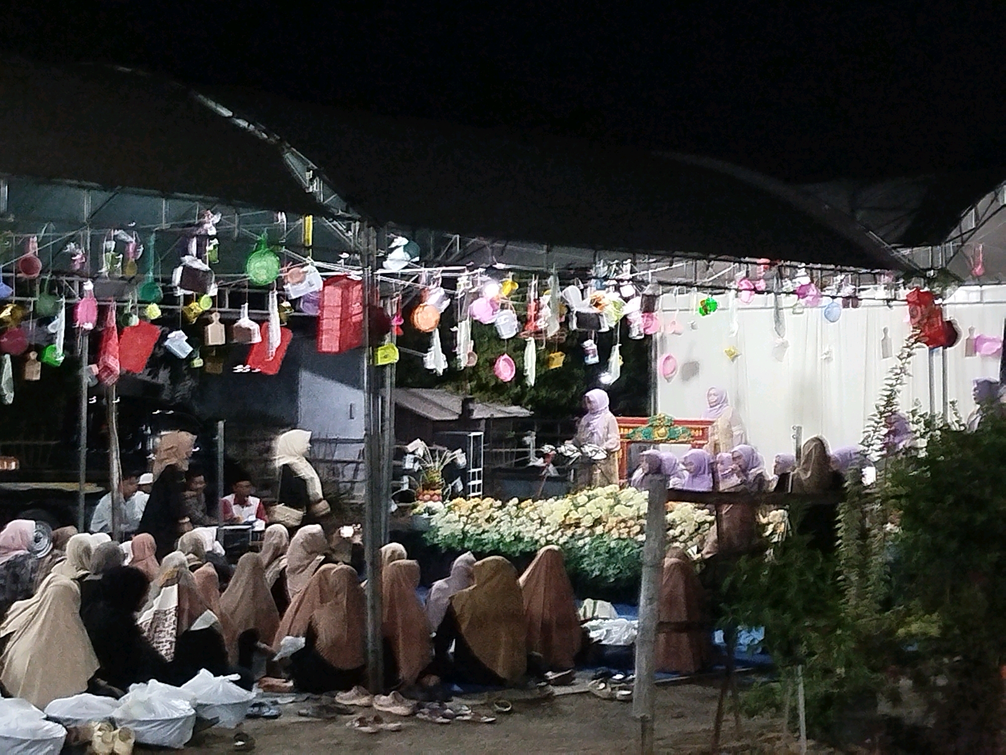 The image depicts an outdoor event at night in Burneh, Indonesia, likely a cultural or religious gathering. A dark canopy covers the scene, supported by metal scaffolding. Hanging from the canopy are numerous colorful decorations, including what appear to be lanterns and small pouches in various shapes and sizes. In the foreground, a group of people, predominantly women wearing hijabs and modest attire, are seated on the ground. Some are interacting, while others appear to be observing. In the midground, a stage or raised platform is visible, adorned with white and yellow floral arrangements. Several individuals, including women in hijabs, are standing on or near the platform. The background features some buildings and trees, suggesting a semi-rural or suburban setting. The overall atmosphere is festive and communal, illuminated by artificial lighting.