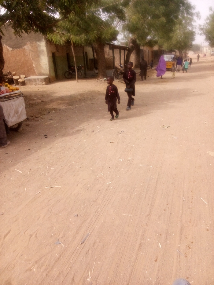 A dusty, sandy road scene features two young boys in matching dark clothing walking away from the camera. Other figures, including one draped in a purple cloth, walk in the same direction. Buildings and trees line the side of the road, and a cart laden with fruit stands to the left. The area suggests a rural or underdeveloped setting. Tire tracks and scattered debris mark the unpaved road.