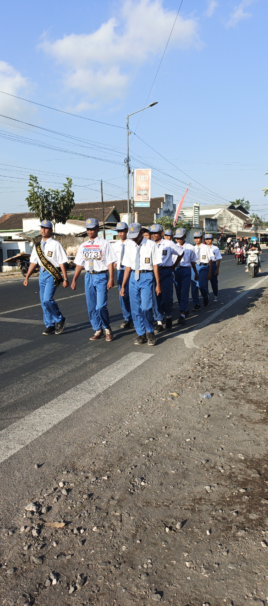 A group of approximately ten male teenagers wearing identical blue trousers, white shirts, grey hats, and grey ties are marching in formation along the right lane of a paved road. Some are wearing blue sashes. The lead individual on the left wears a black sash with gold lettering that reads "SMA NEGERI KASIYAN". Another participant in the front row has a white bib with the number "073" in red. The teenagers are marching in a procession, likely for a school event or parade. The road is bordered by a shoulder of dirt and gravel, with some small rocks and debris visible. In the background, there are houses with tiled roofs, a street lamp, power lines, and a few people on motorcycles on the opposite side of the road. A red and white flag is visible on a pole in the middle ground. The sky is clear blue with a few scattered clouds. The scene appears to be in Kasiyan, Indonesia, as indicated by the sash. The overall atmosphere suggests a daytime outdoor event under sunny conditions.