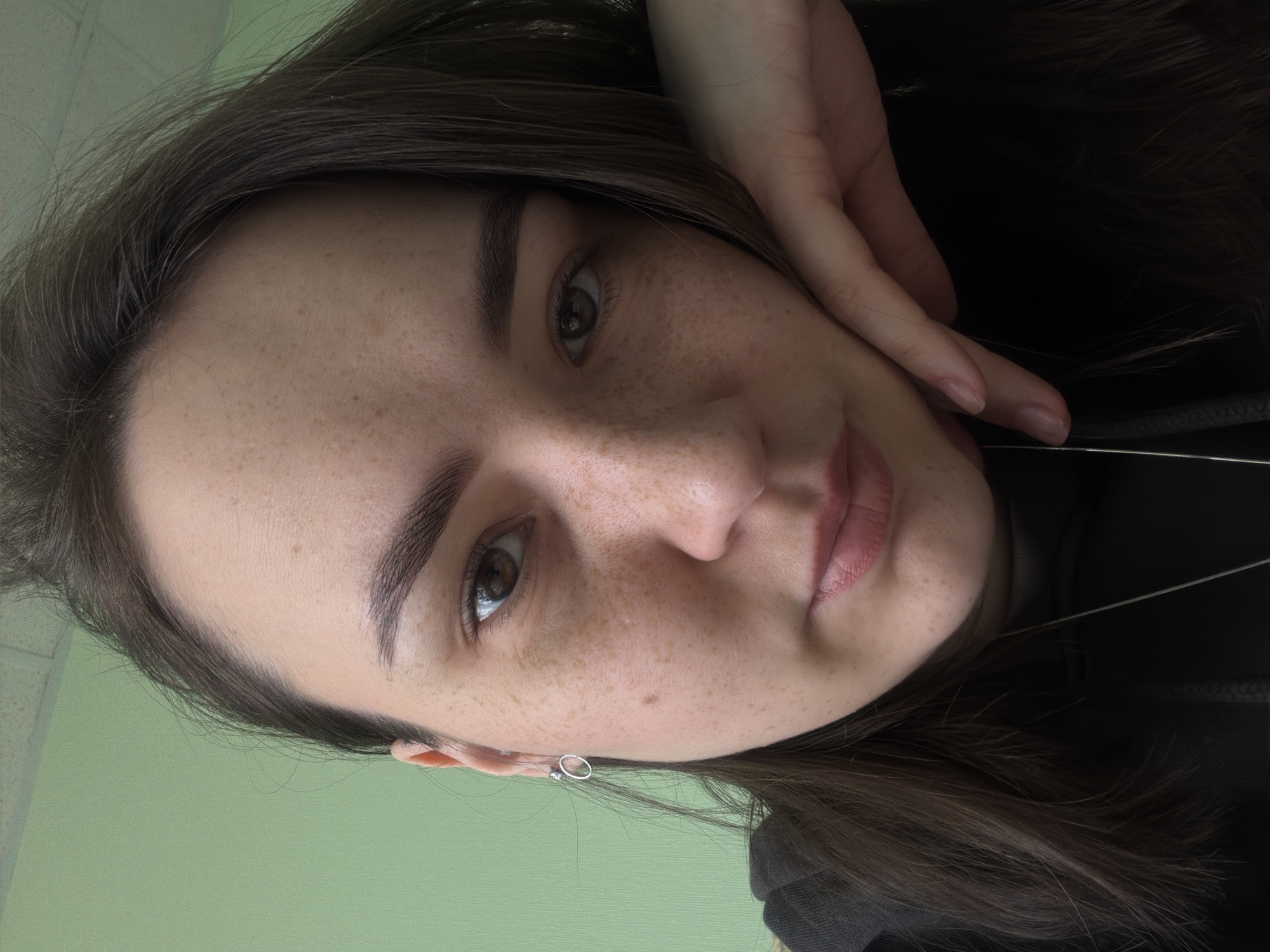 A close-up, tilted portrait captures a young person, likely female, taking a selfie. The subject has medium-long, dark brown hair, prominent dark eyebrows, and warm brown eyes that gaze directly at the camera. Their fair skin features numerous visible freckles across the forehead, nose, and cheeks, suggesting a natural complexion. A subtle, soft smile plays on their lips, which have a natural pink hue. One hand, with neatly kept nails, is gently resting near the side of their face and hair on the right, partially obscuring the background.
The person is wearing a dark-colored top, possibly a hoodie or sweatshirt, indicated by the material and a visible drawstring. A delicate silver-toned necklace is worn around the neck, and a small silver hoop earring, along with another tiny stud, is visible in their left earlobe.
The background primarily consists of a plain, light green wall visible on the left side of the frame, providing a soft contrast to the dark hair. The lighting is soft and even, suggesting an indoor setting. The overall mood conveyed is one of calm and natural simplicity. The image was taken in Chaikovsky, Russia. There is no visible text.