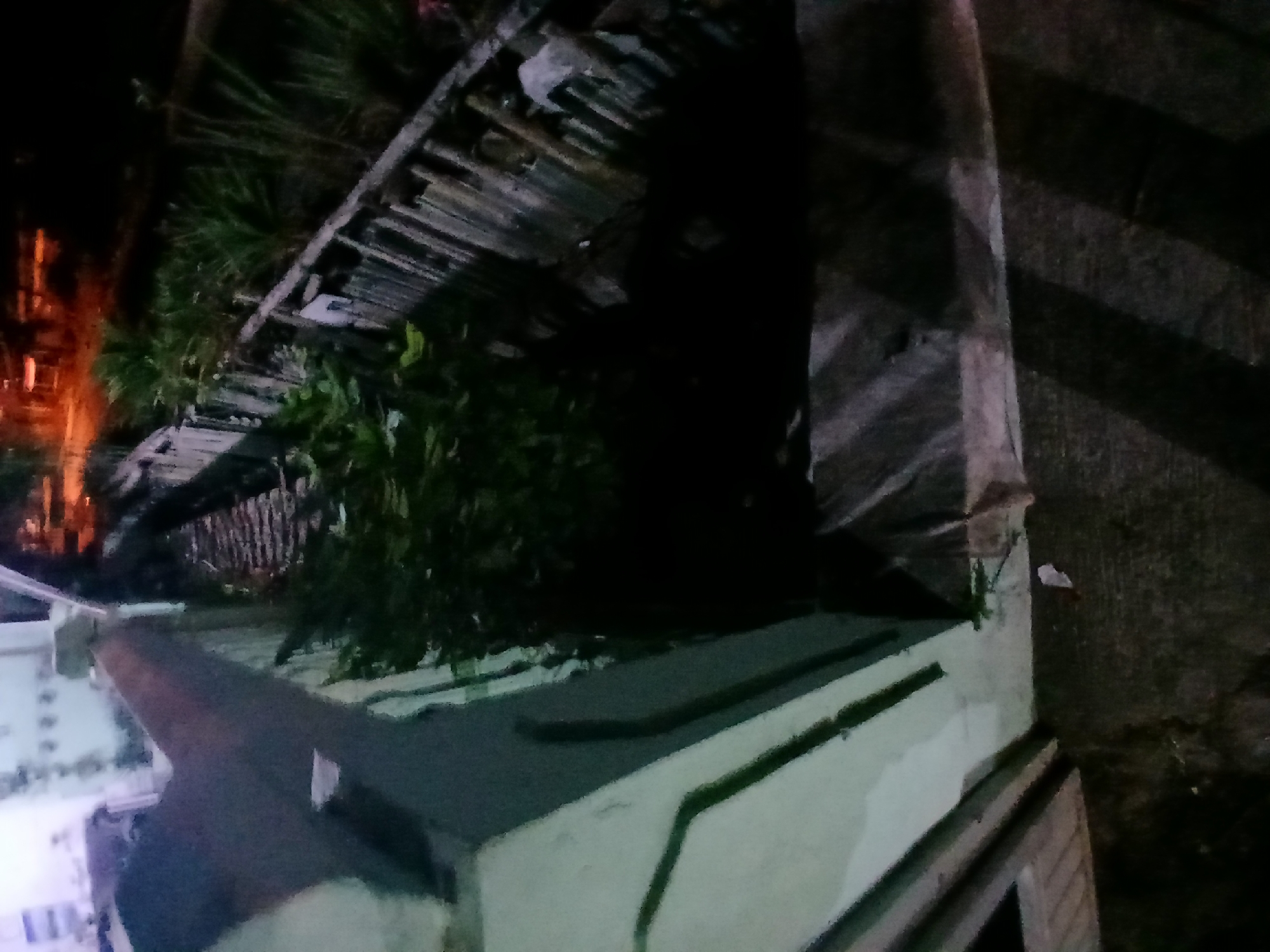 The image depicts an outdoor scene at night. In the foreground, a white wall with green trim runs diagonally from the left to the right. A dark, angular structure is visible in the upper left quadrant, casting a shadow. Beyond the wall, a wooden fence with corrugated metal sheeting is visible, lined with dense green foliage. The background is dark with some orange lights suggesting distant buildings or streetlights. The ground appears to be paved or covered in concrete. There are no people or discernible actions taking place. The scene is consistent with a residential or industrial area in Kademangan Kulon, Indonesia, at night.