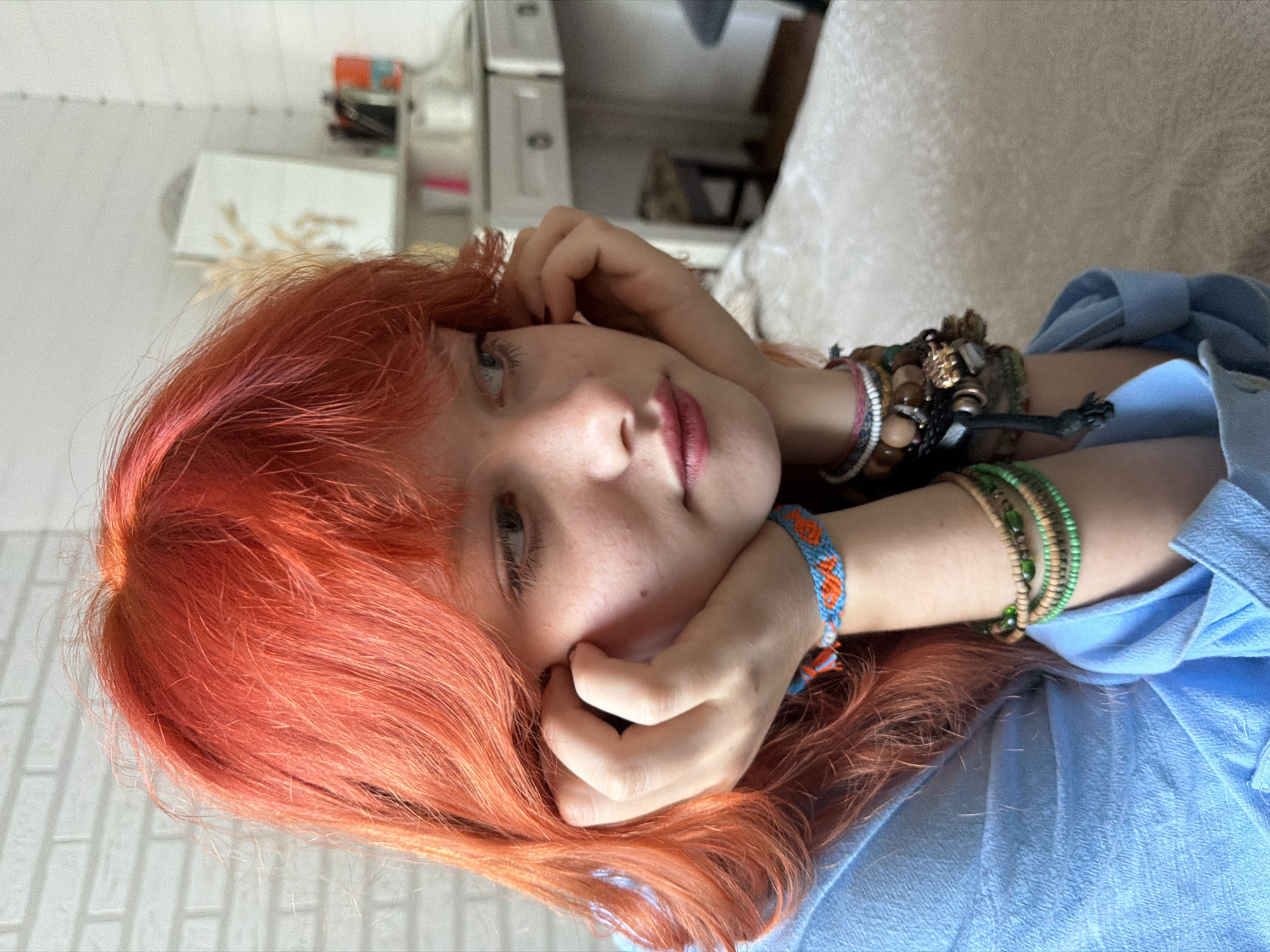 The media file shows a close-up, portrait-oriented image of a young person with bright orange hair lying down. Their head is tilted, and one hand is resting on their cheek, while the other arm is bent with bracelets visible on their wrist. They are wearing a light blue collared shirt. The background is slightly blurred, showing what appears to be an indoor setting with furniture, including what might be a dresser or shelves with some objects on them, and a patterned wall. The lighting suggests it is daytime. The person's gaze is directed slightly off-camera, and their expression appears relaxed or contemplative. There is no visible text in the image. The provided location context is Sochi, Russia.