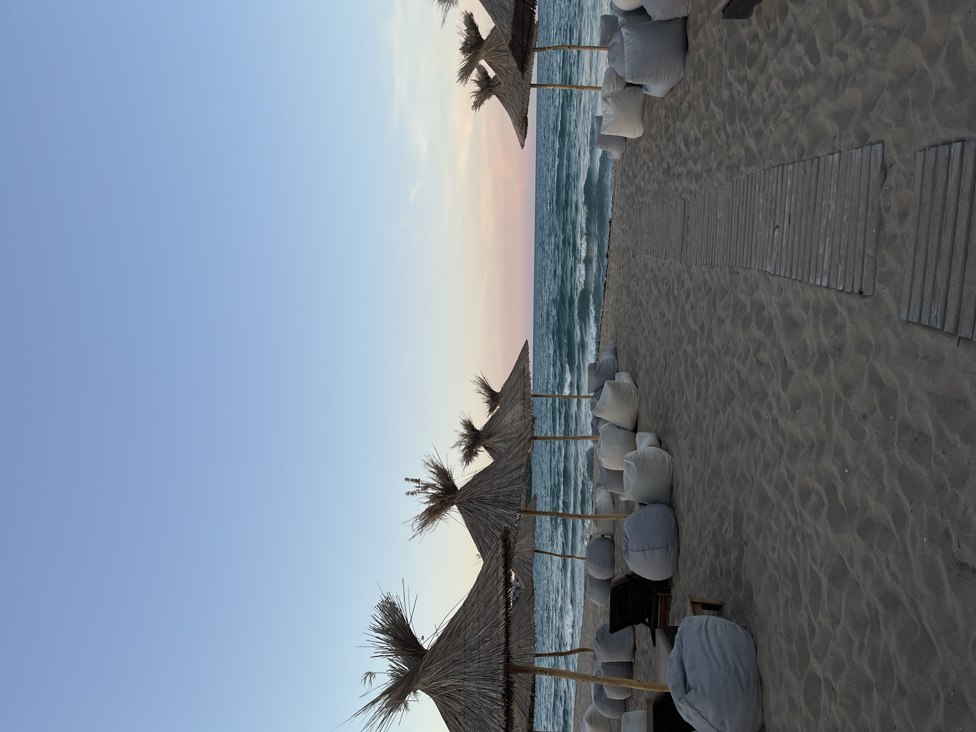 A beach scene in Sozopol, Bulgaria, at dusk. The sky is a gradient of pale blue to soft pink. The sea is a deep blue with gentle waves. On the sandy beach, there are several straw umbrellas providing shade. In front of the umbrellas, there are many large, white and grey beanbag chairs and a few dark-colored lounge chairs. A wooden walkway runs through the sand. There are no people visible in the image.