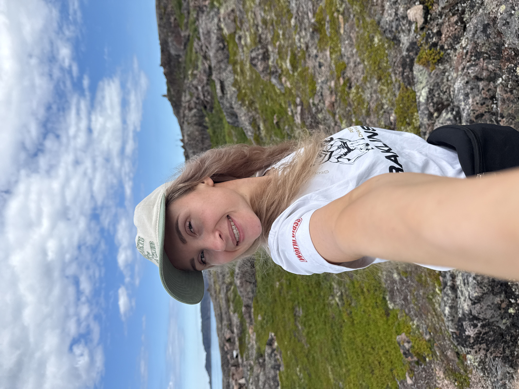A young woman with blonde hair and braces is taking a selfie outdoors in a rocky, green landscape with a body of water in the background. She is wearing a white t-shirt with a graphic on it and a light-colored baseball cap. The sky above is blue with white clouds. The scene appears to be in Teriberka, Russia, given the characteristic rocky terrain and the proximity to the Arctic Ocean.