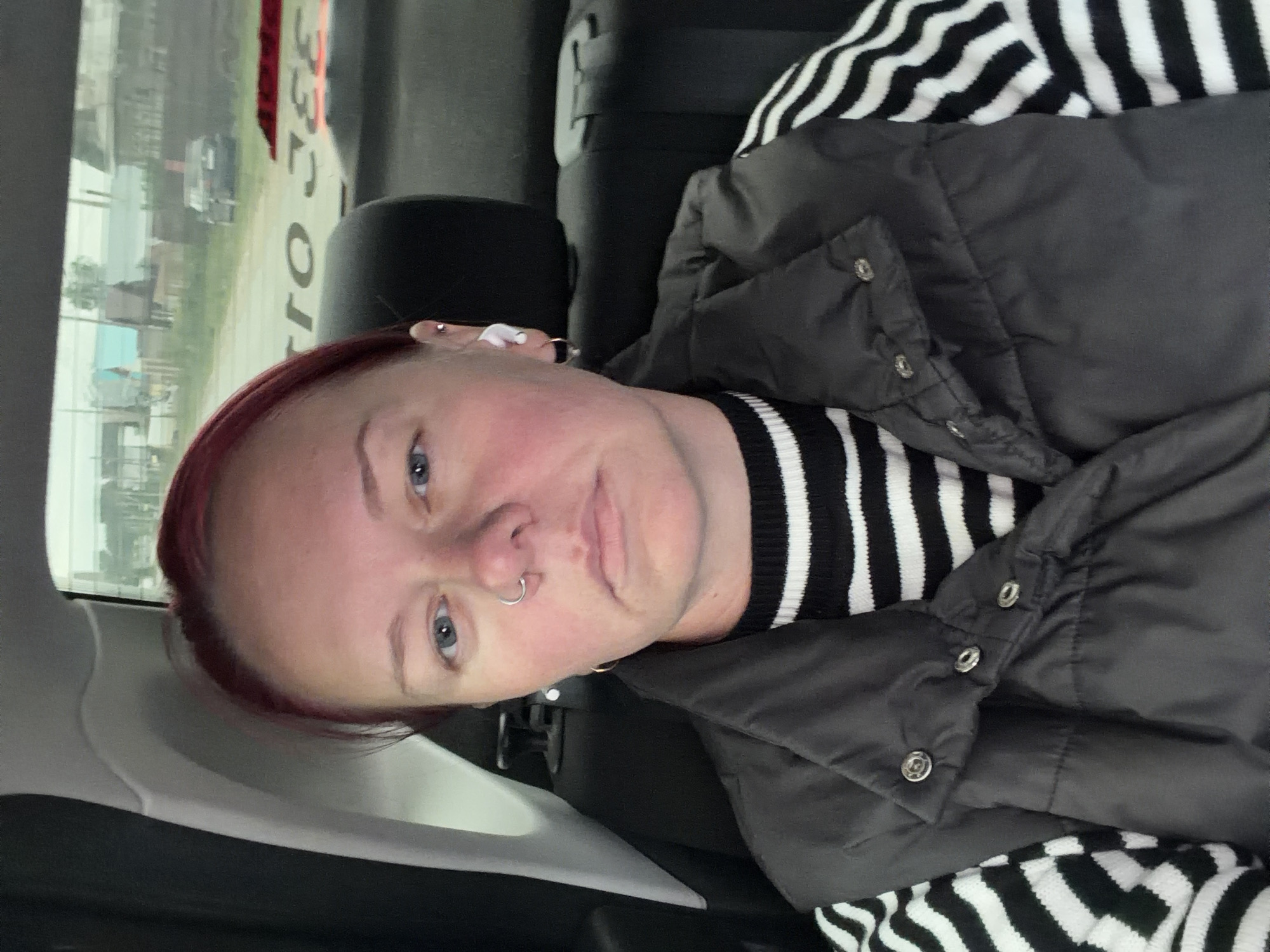 A woman with red hair and blue eyes is in the back seat of a car, looking at the camera. She is wearing a black vest over a black and white striped sweater. She has a nose ring and an ear piercing. The background shows a blurry view of a village, likely in Chaikovsky, Russia, with houses and trees. There is text on the back window of the car that is partially visible.