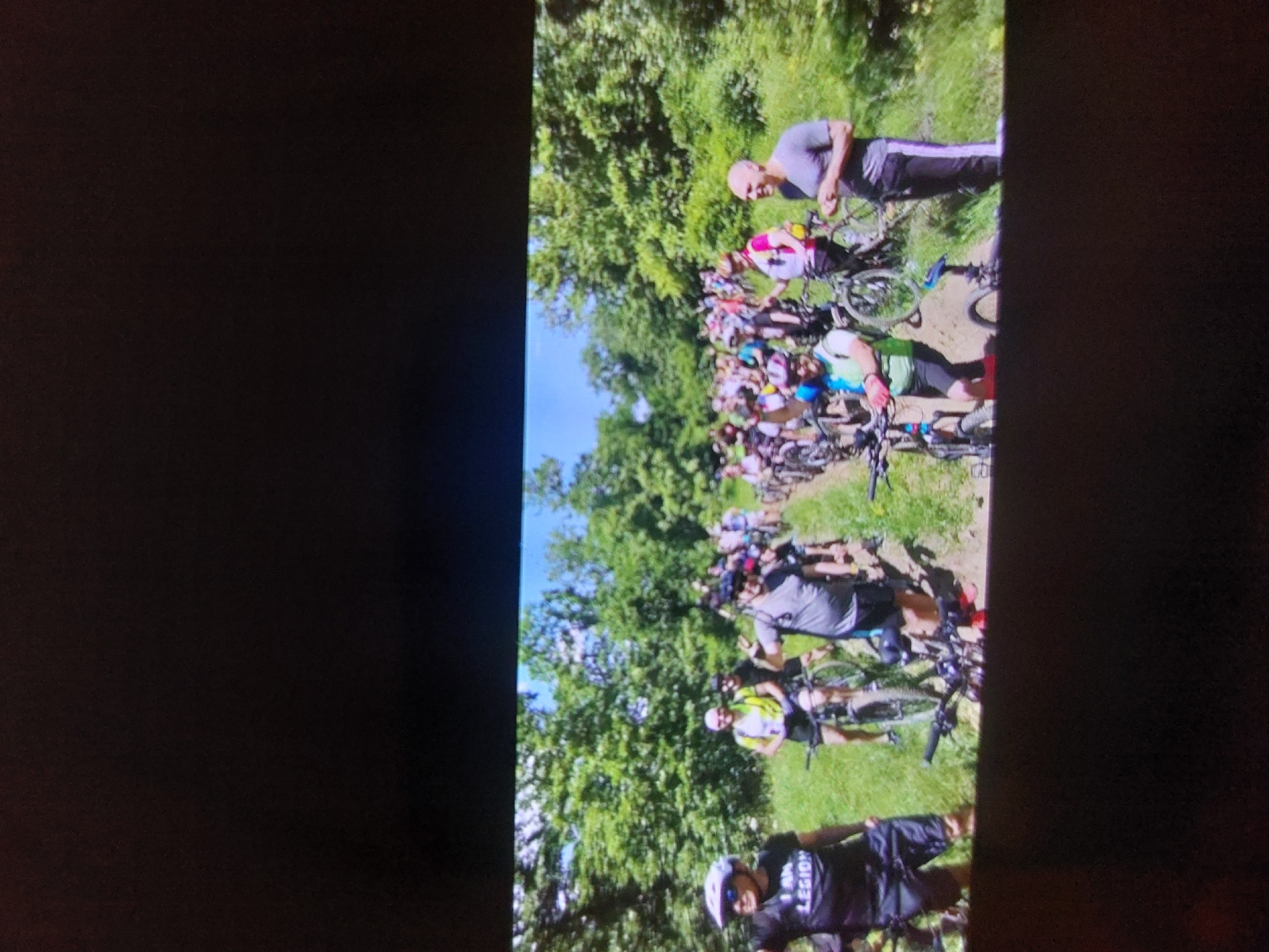 The image captures a vibrant outdoor gathering of numerous mountain bikers, set against a backdrop of lush green trees and a clear blue sky. The photograph is presented with a 90-degree counter-clockwise rotation, so the main activity unfolds along a vertical axis.
The subjects are a large group of people, primarily adults, engaged in what appears to be a mountain biking event or group ride. They are dressed in typical cycling attire, including colorful jerseys, shorts, and safety helmets. Many individuals are standing next to their mountain bikes or are in the process of mounting them.
Objects prominently featured include a variety of mountain bikes, each equipped for off-road terrain, with visible handlebars, frames, and wheels. Helmets are worn by most participants, indicating a focus on safety. The natural environment is dominated by dense green foliage, tall trees, and a visible dirt path or trail where the group is assembled.
The scene depicts a collective activity, likely a start of a group ride, a race, or a social mountain biking excursion. The participants appear to be paused, suggesting they are waiting for instructions, taking a break, or preparing to set off.
Notable details include the bright, sunny weather and a clear blue sky, indicating a pleasant day for outdoor activity, likely late morning or early afternoon. The vibrant green of the foliage suggests a spring or summer season. The general atmosphere seems positive, with some individuals smiling towards the camera, conveying a sense of camaraderie and enjoyment. A distinct piece of text is visible on a black jersey worn by a person in the foreground (bottom left when viewed upright), clearly stating "I AM LEGION". The setting, with its forested trails, aligns well with the provided context of Tsarevo, Bulgaria, which offers suitable terrain for mountain biking.