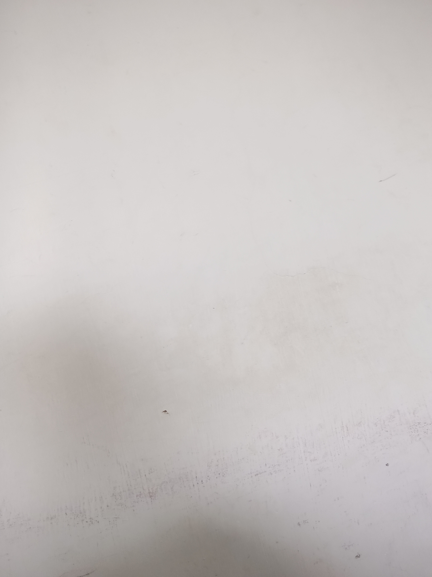 The image shows a close-up view of a pale, textured surface, possibly a wall or a piece of furniture, with subtle variations in shading and some minor imperfections. There are faint smudges and a few small, dark specks scattered across the surface. A small, light-colored imperfection, possibly a chip or scratch, is visible in the lower-left quadrant of the image. The overall impression is one of a plain, perhaps aged, surface. The image does not contain any identifiable elements that would suggest its location in Lhokseumawe, Indonesia, or any specific environmental context.