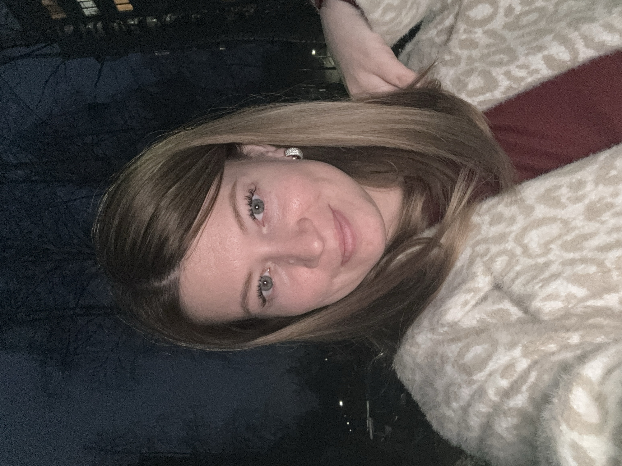A woman with light brown hair and green eyes is posing for a portrait in an outdoor setting. She is wearing a patterned, light-colored coat. The background appears dark, possibly taken at night, with a suggestion of trees and distant city lights, hinting at an urban environment. There's no specific information available within the image to confirm a location in Chaikovsky, Russia.
